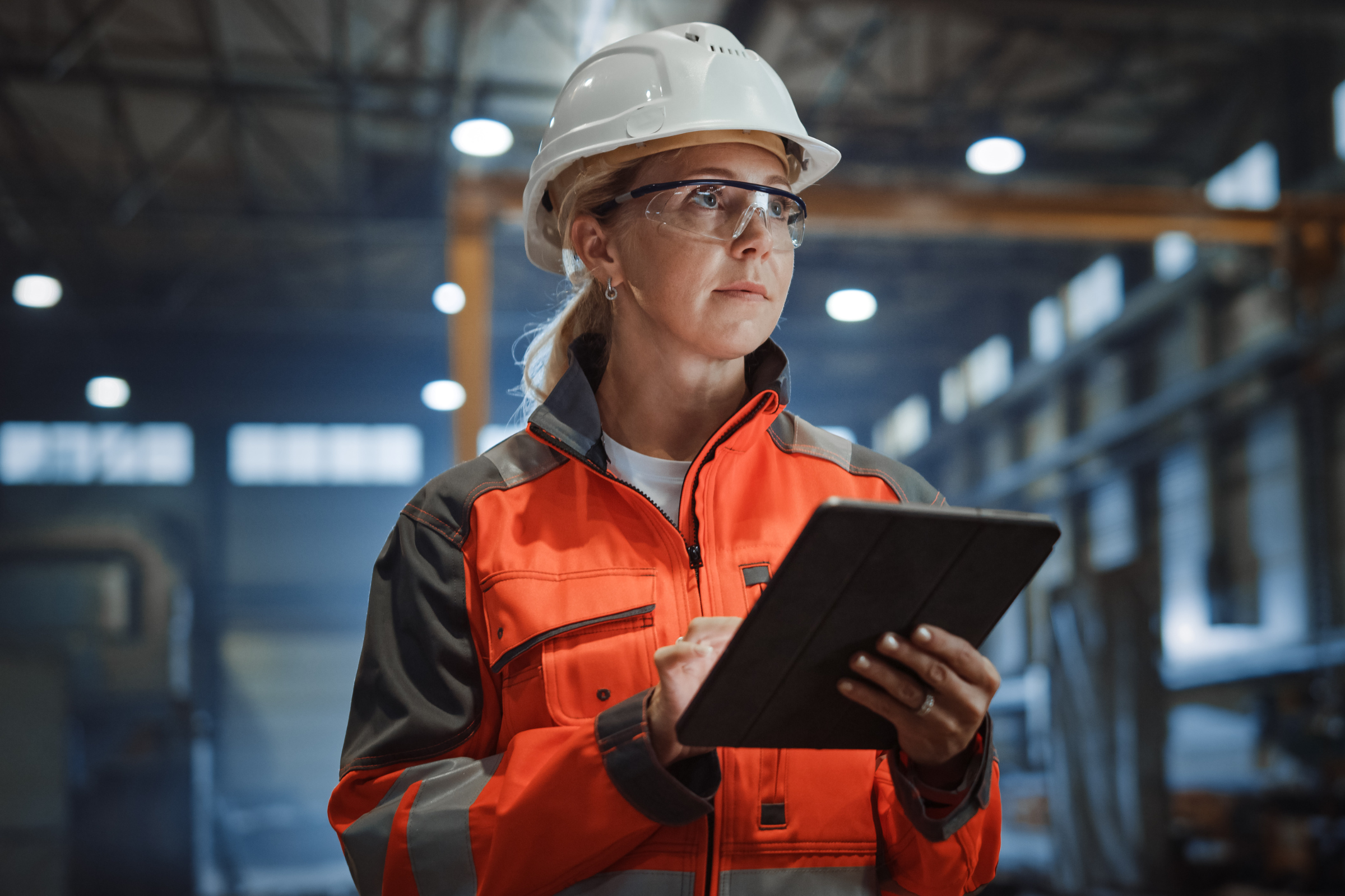 Woman engineer wearing safety helmet and glasses holding a tablet in an industrial warehouse.