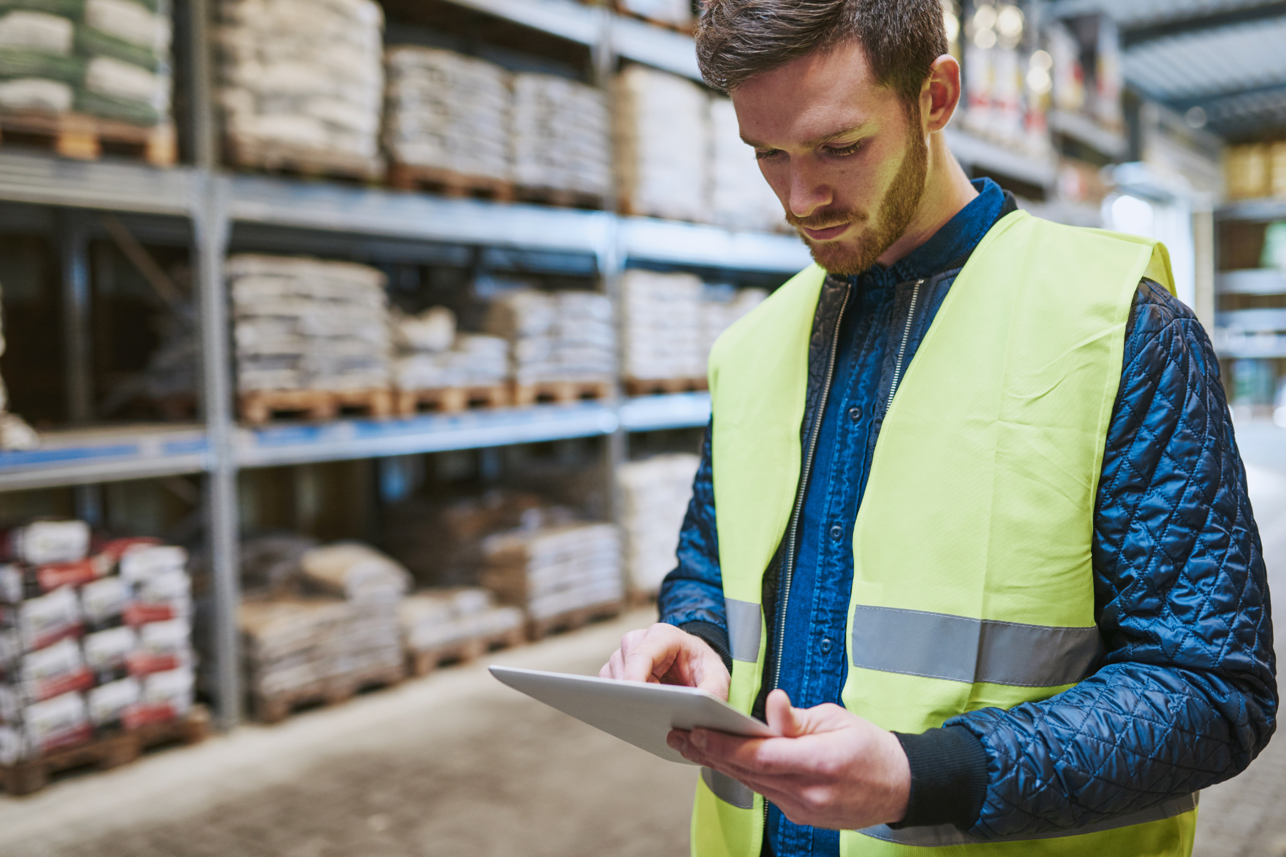 Man in a high-visibility vest using a tablet in a warehouse with shelves stacked with goods in the background.