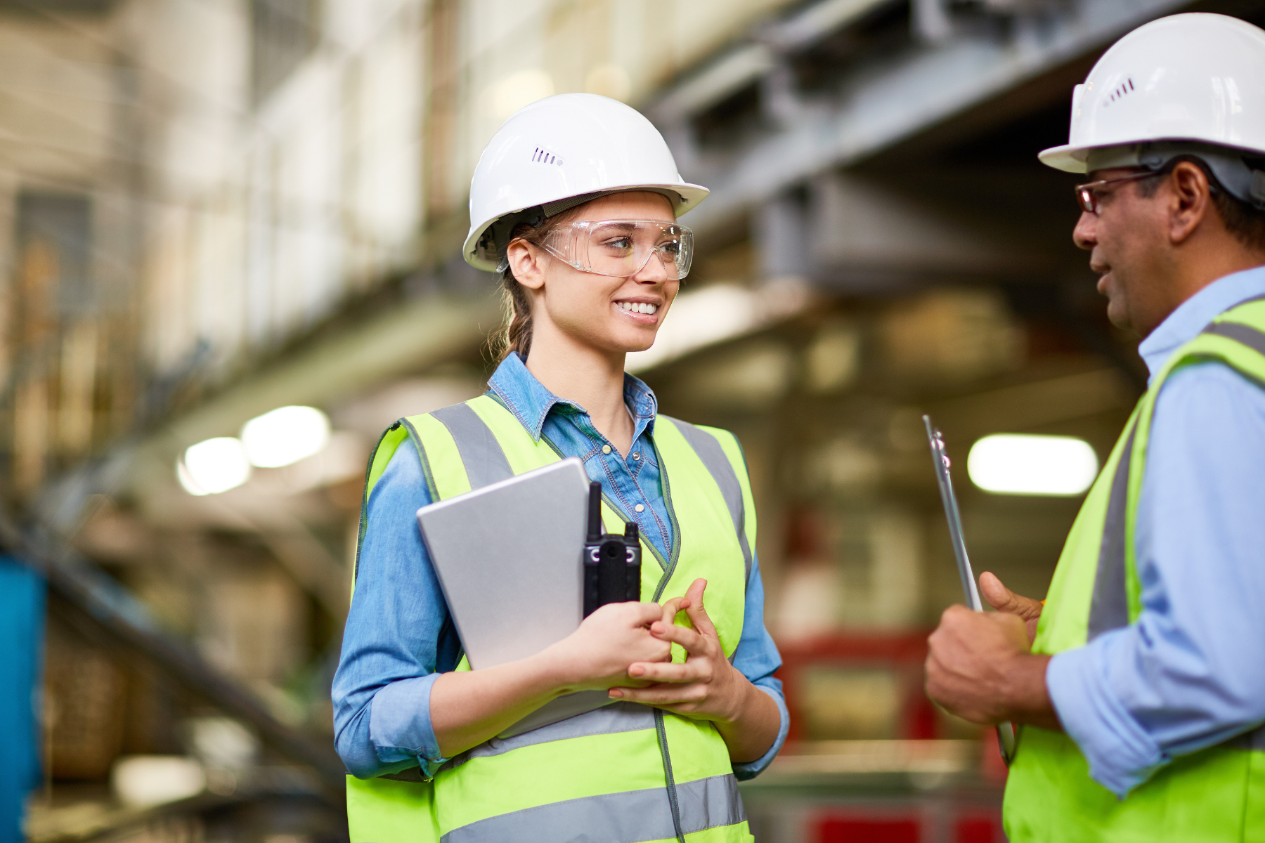 Two factory workers wearing hard hats and safety vests having a discussion in an industrial setting.