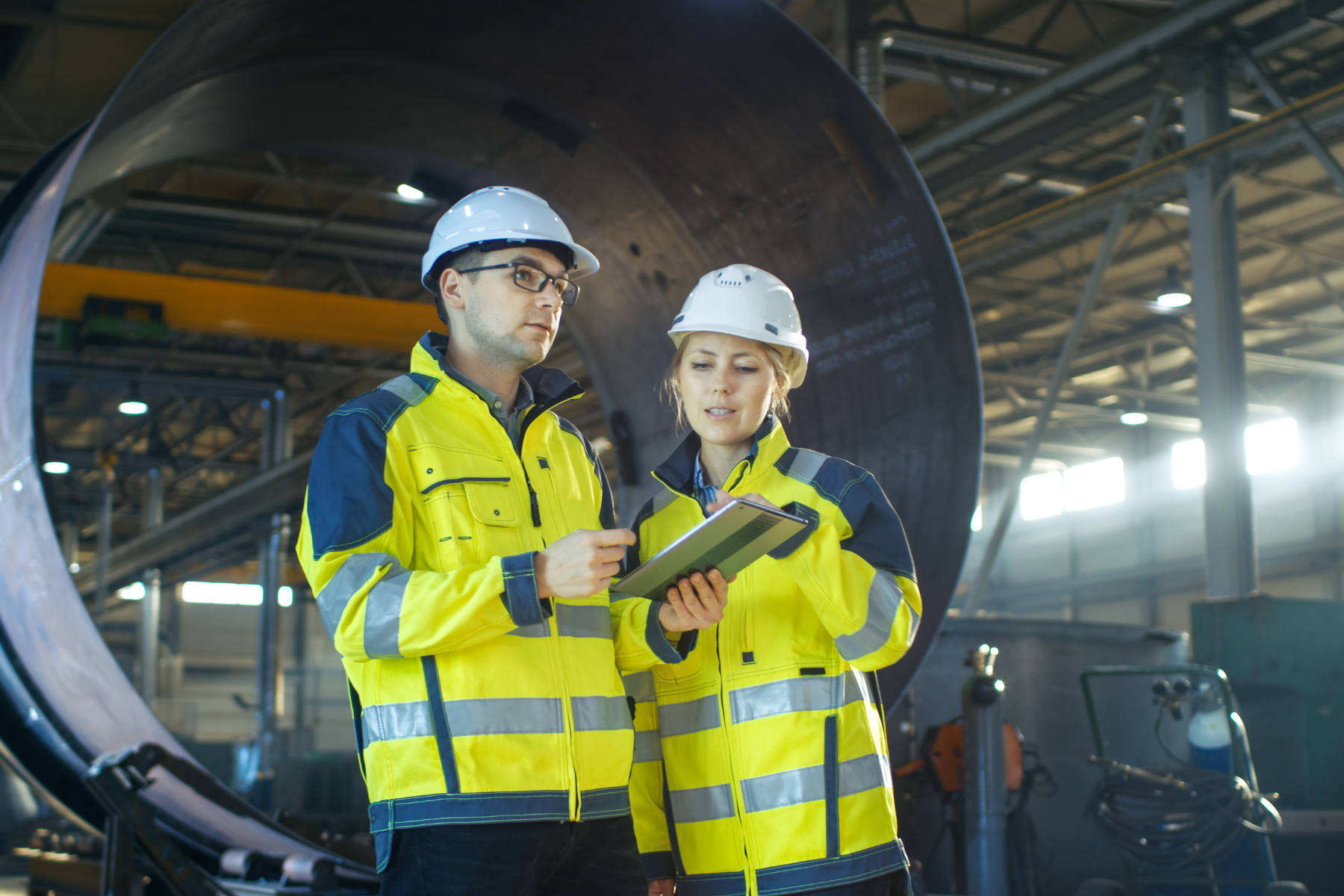Two engineers in yellow reflective jackets and white helmets reviewing a tablet inside an industrial facility.