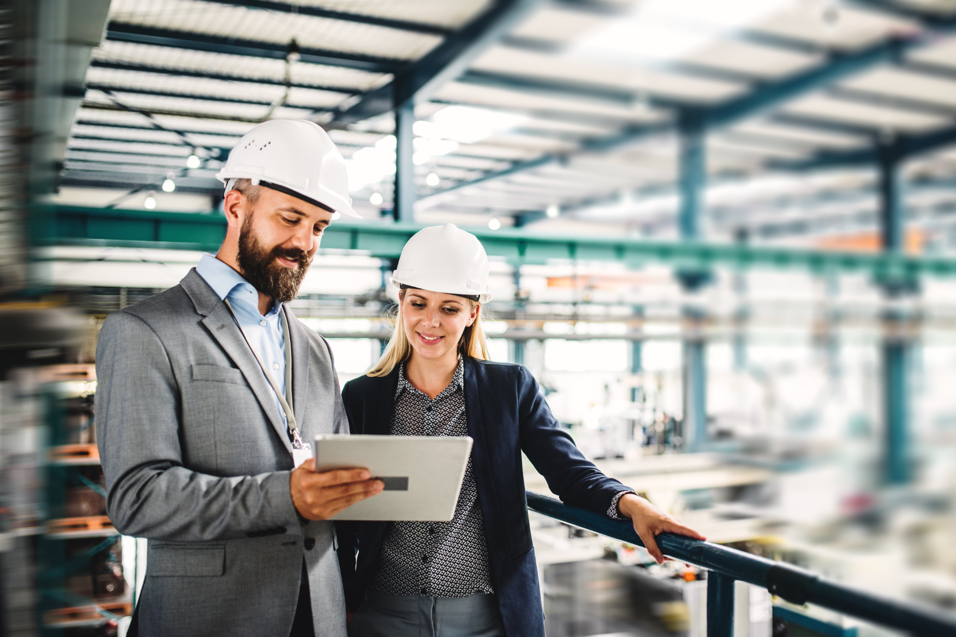Two engineers wearing white hard hats are reviewing information on a tablet in an industrial facility.