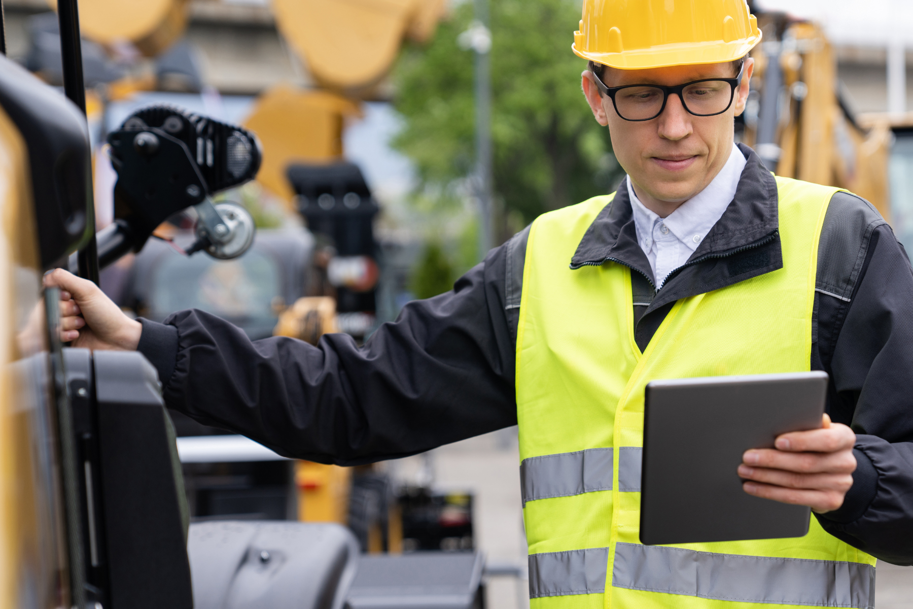 Construction worker wearing a yellow safety vest and hard hat using a digital tablet while standing next to heavy machinery.
