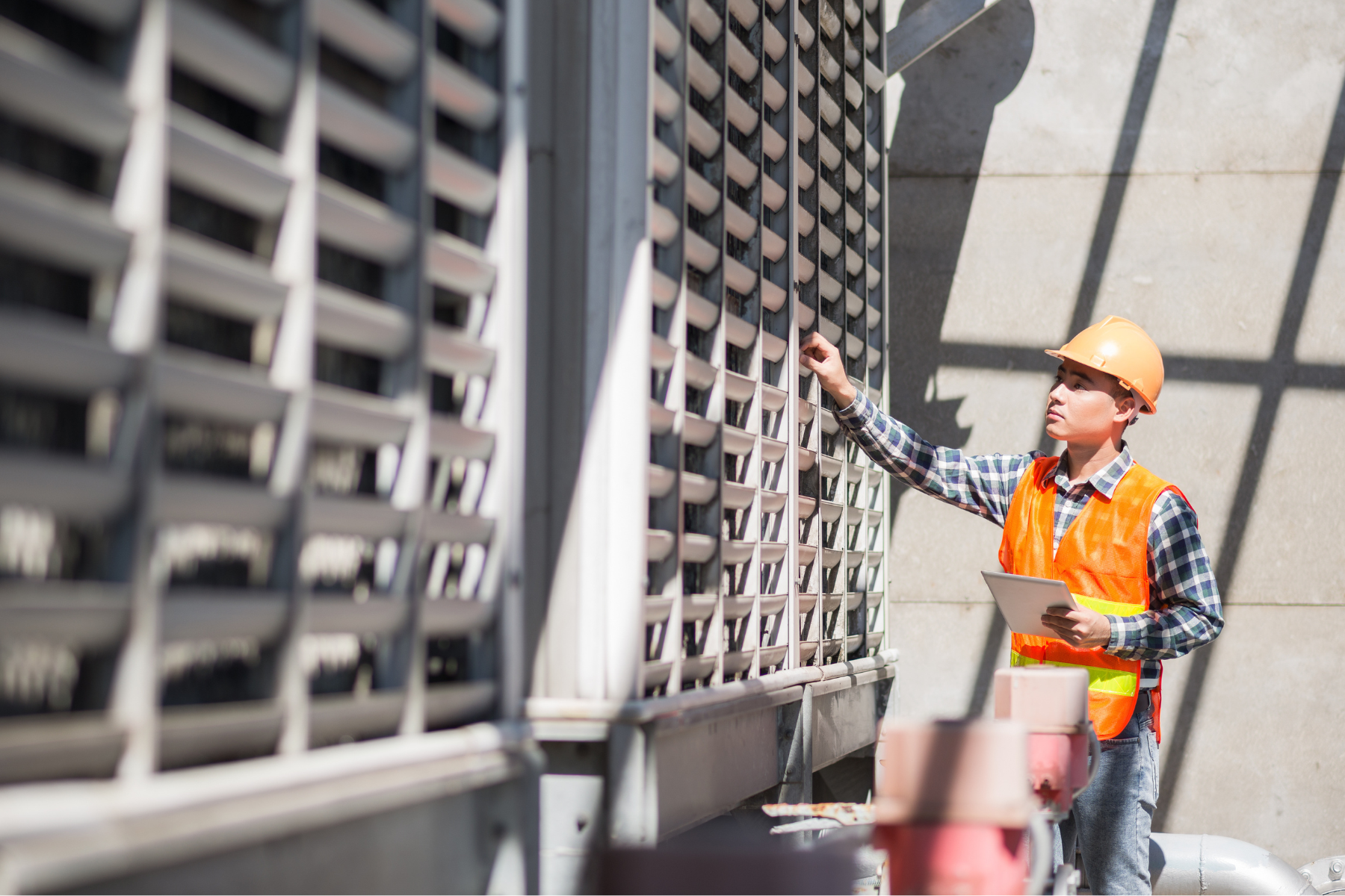 Construction worker wearing an orange safety vest and hard hat inspecting industrial machinery while holding a tablet.