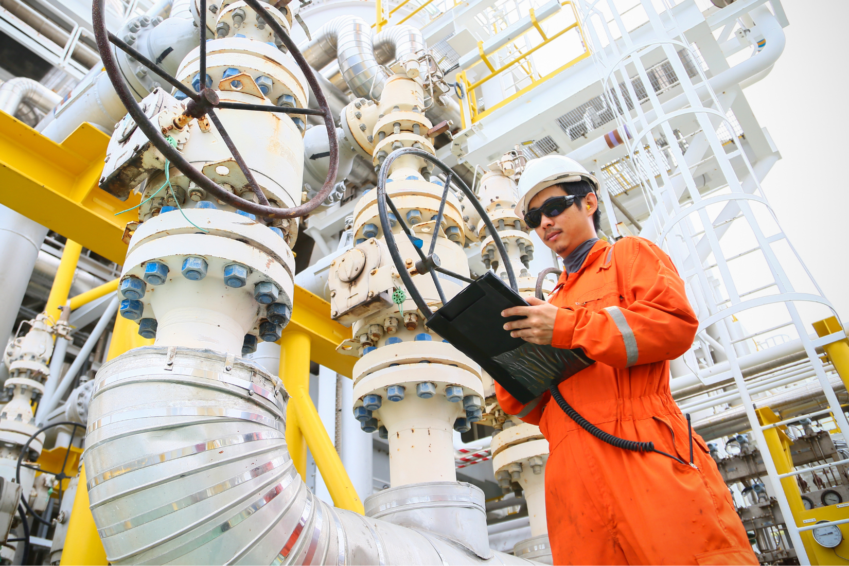 Worker in orange safety coverall and white helmet using a tablet while inspecting equipment in an industrial plant.