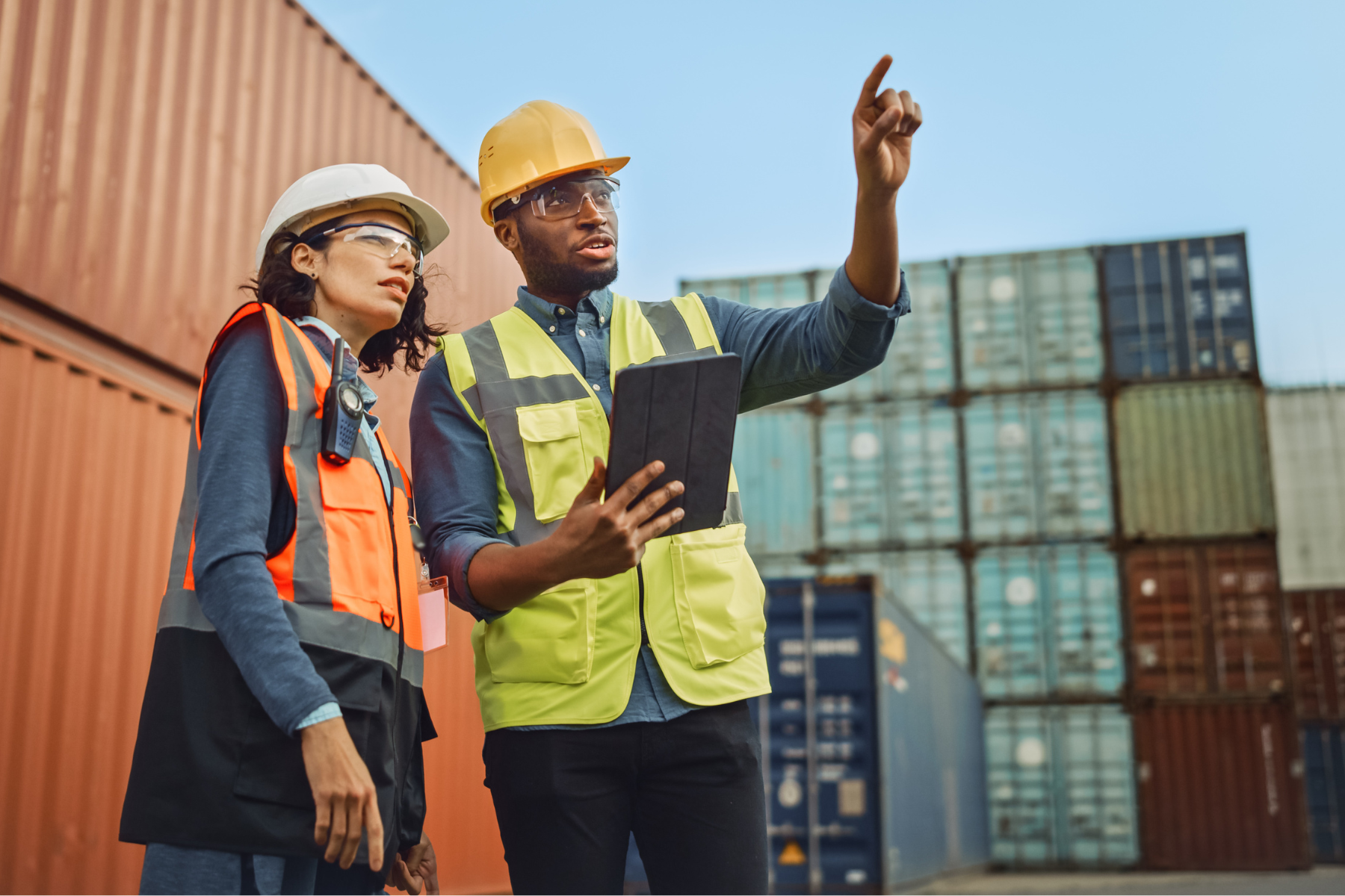Two logistics workers wearing safety helmets and vests inspecting shipping containers, one holding a tablet and pointing.