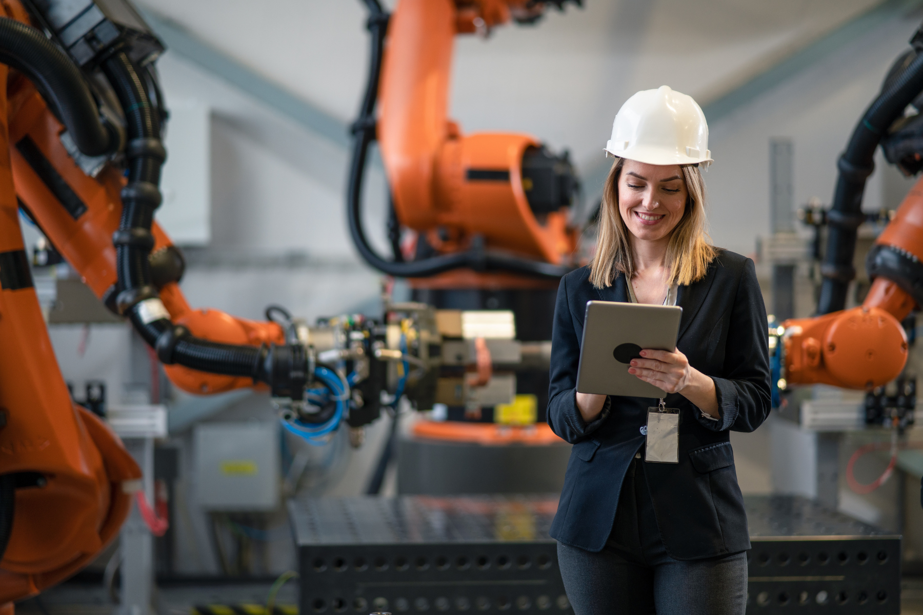 Woman in a white hard hat using a tablet while standing in an industrial setting with orange robotic arms.