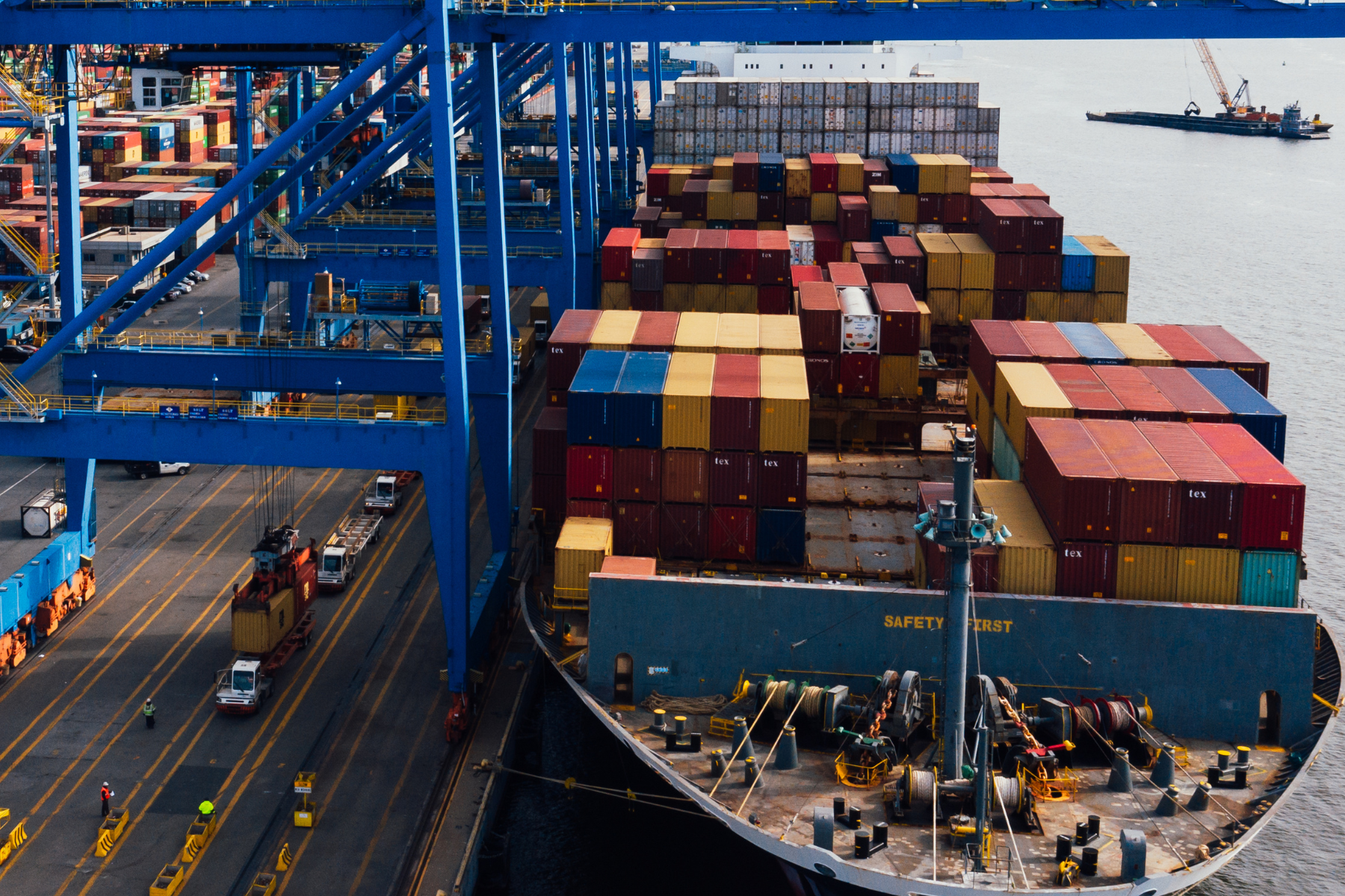 Cargo ship loaded with multicolored shipping containers docked at a port with large blue cranes unloading containers.