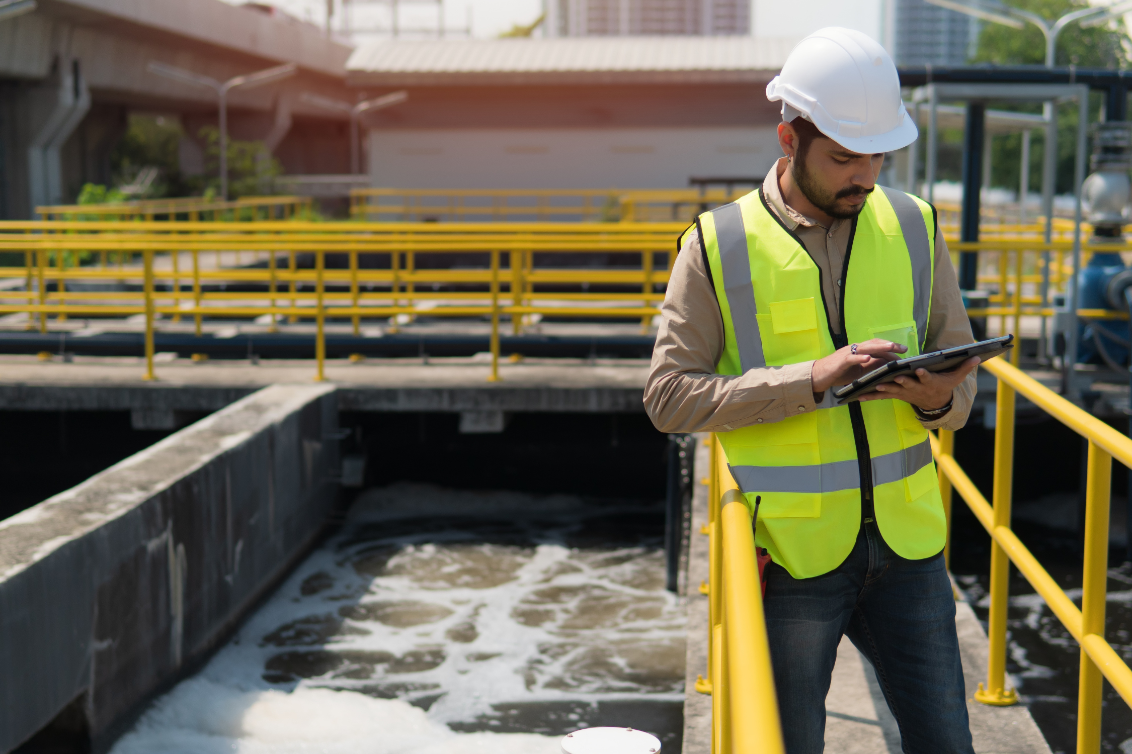 Male engineer in a white hard hat and yellow reflective vest using a tablet at a water treatment facility.