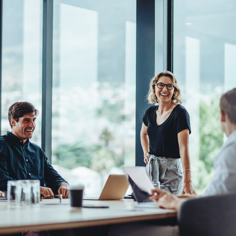 Three colleagues in a bright office engaged in a lively discussion around a table with laptops and documents.