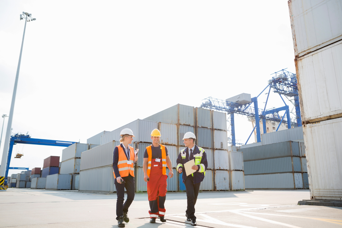 Three workers wearing safety helmets and reflective vests walking in a shipping container yard with stacked containers and cranes in the background.