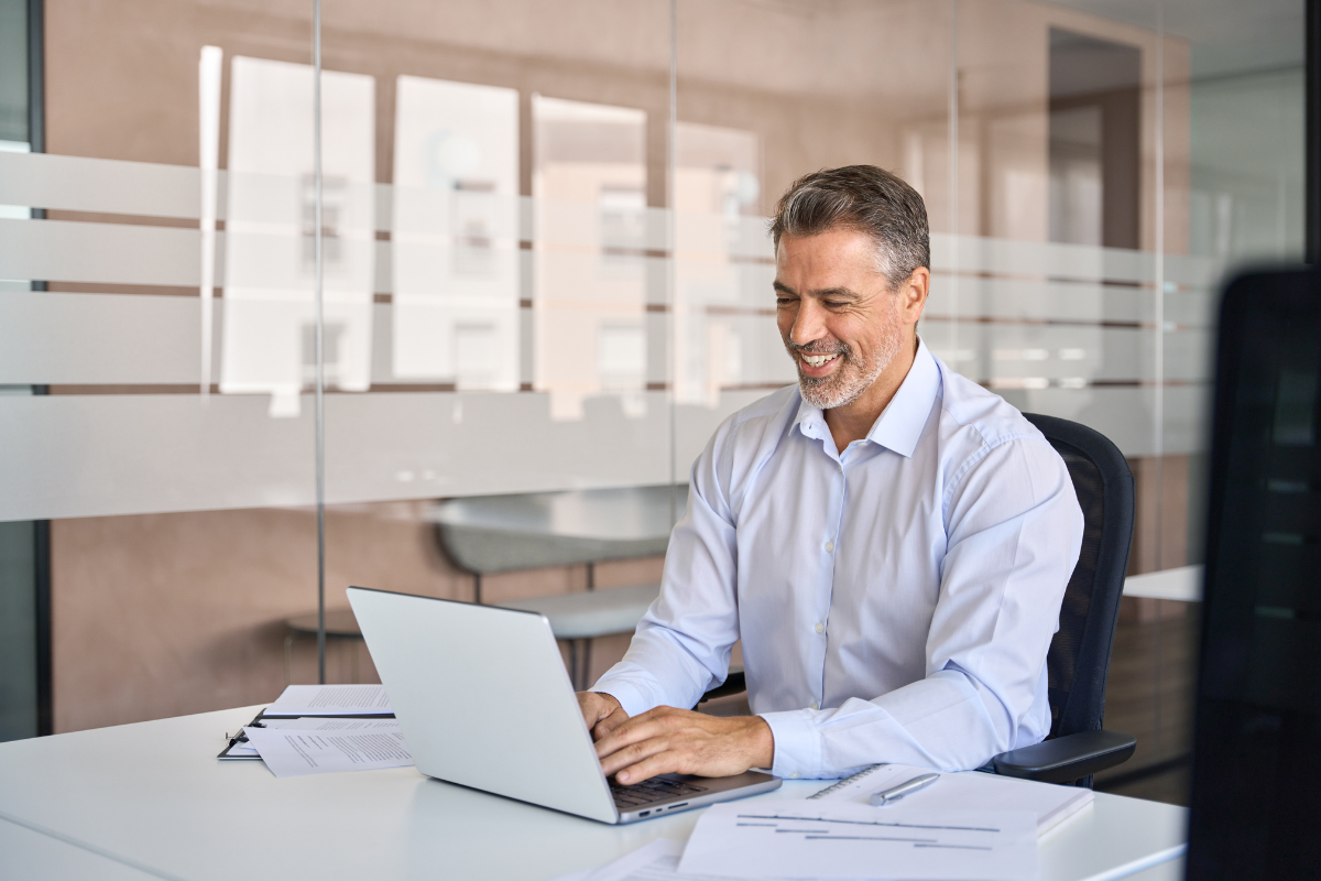 Smiling man in white shirt working on a laptop at a white desk in a modern office.