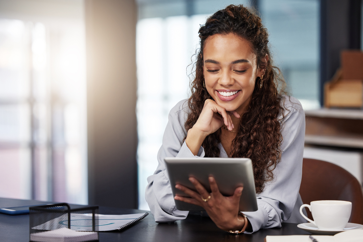 Smiling young woman with curly hair using a tablet at a desk with a cup of coffee and notebook nearby.