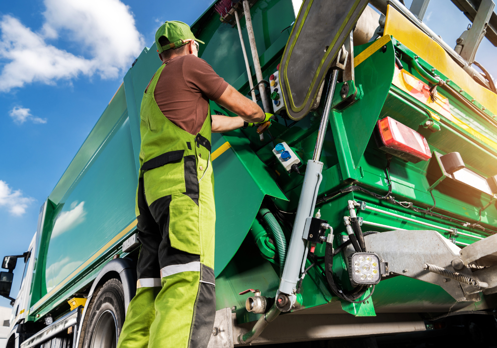 Worker in green overalls operating a green garbage truck under a partly cloudy blue sky.