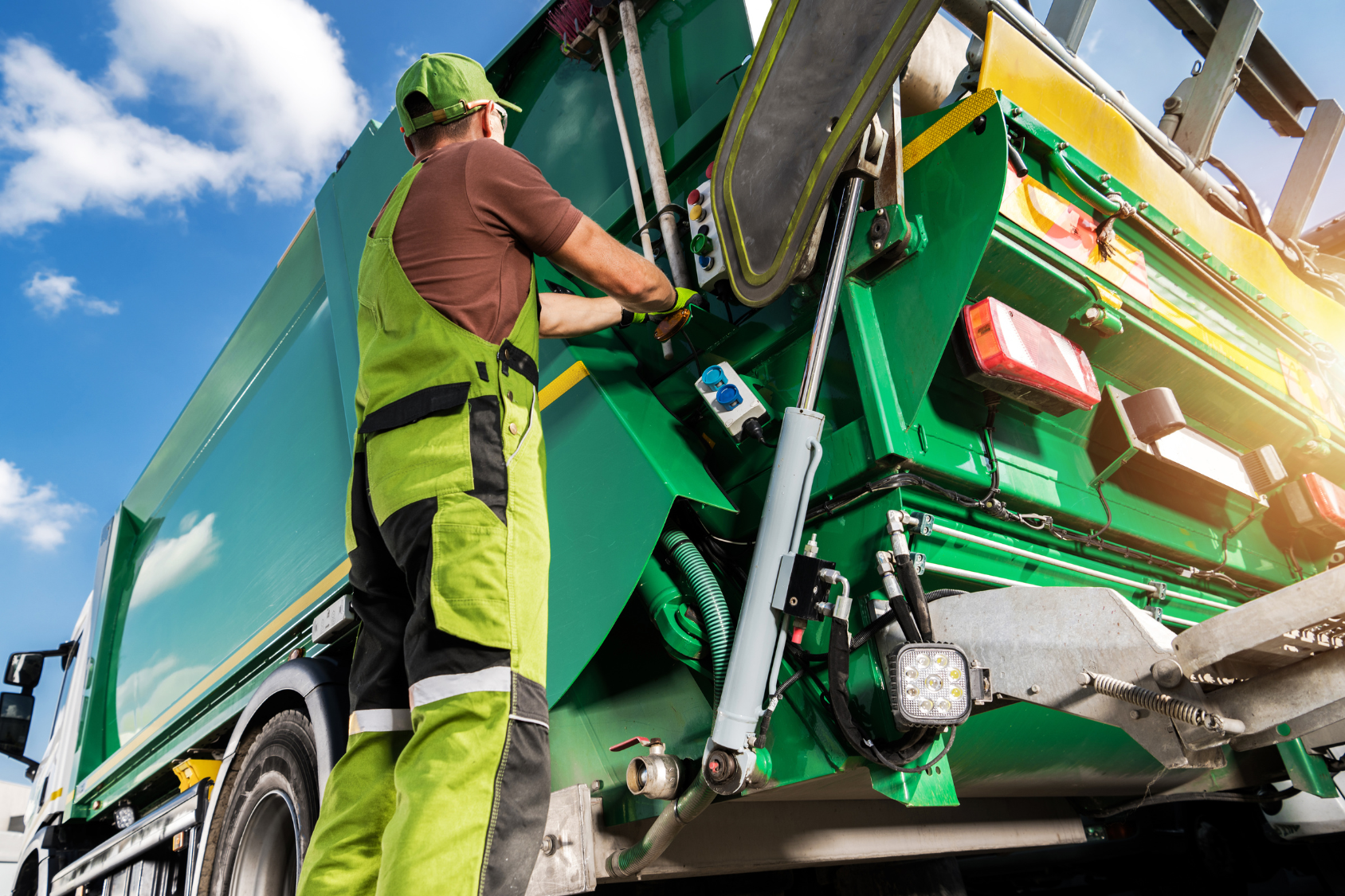 Worker in green and black uniform operating a green waste management truck under a clear blue sky.