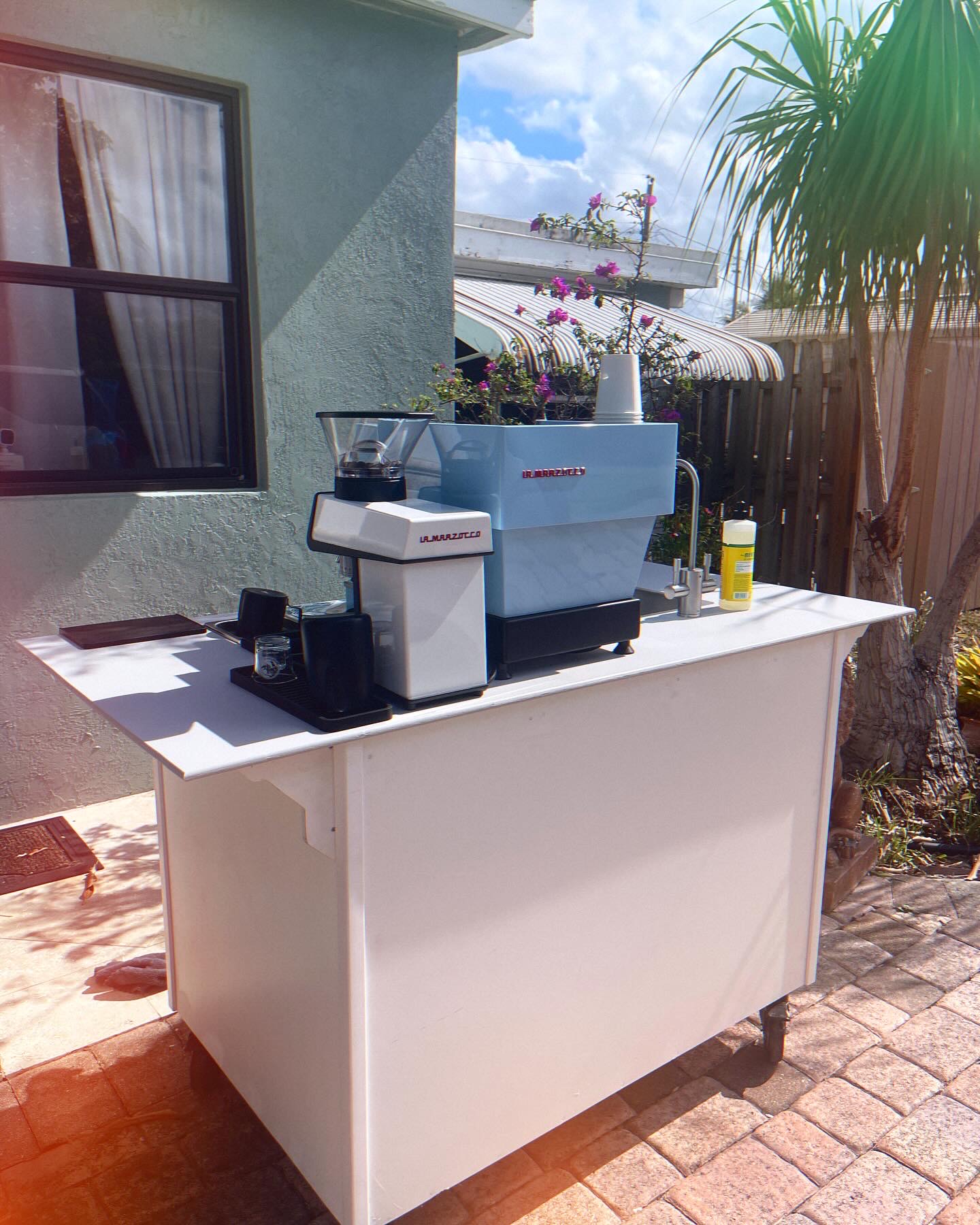 Outdoor portable white coffee bar with La Marzocco espresso machine and grinder on a patio.
