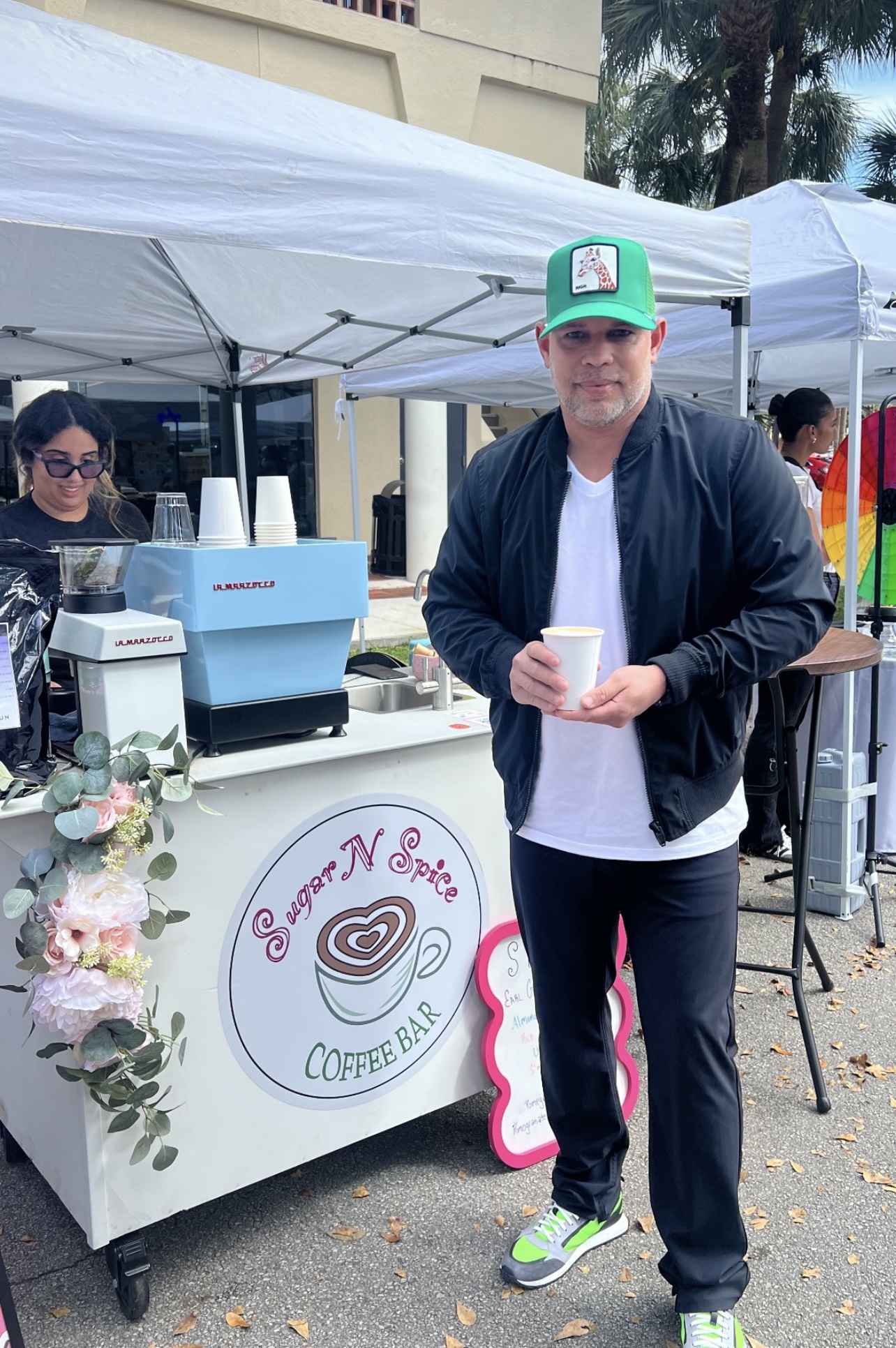 Man wearing green hat holding a coffee cup standing next to Sugar N Spice Coffee Bar stand.