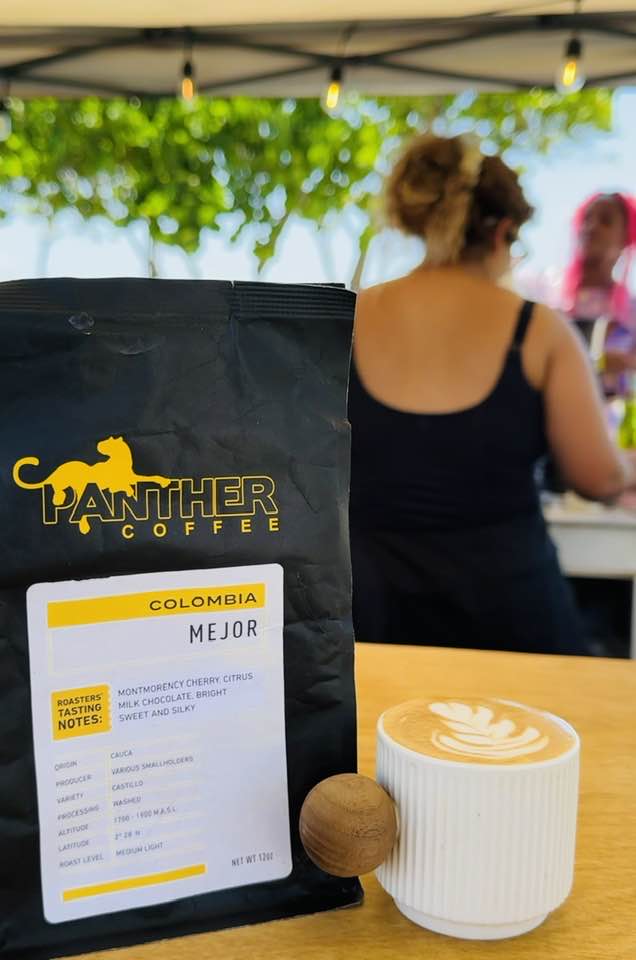Black bag of Panther Coffee Colombia Mejor with tasting notes next to latte with leaf latte art in Boca Raton, FL