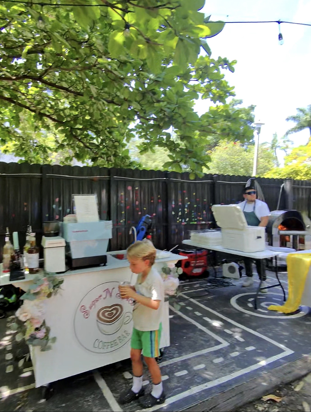 Young boy drinking from a cup beside a coffee bar and adult near an outdoor pizza oven.