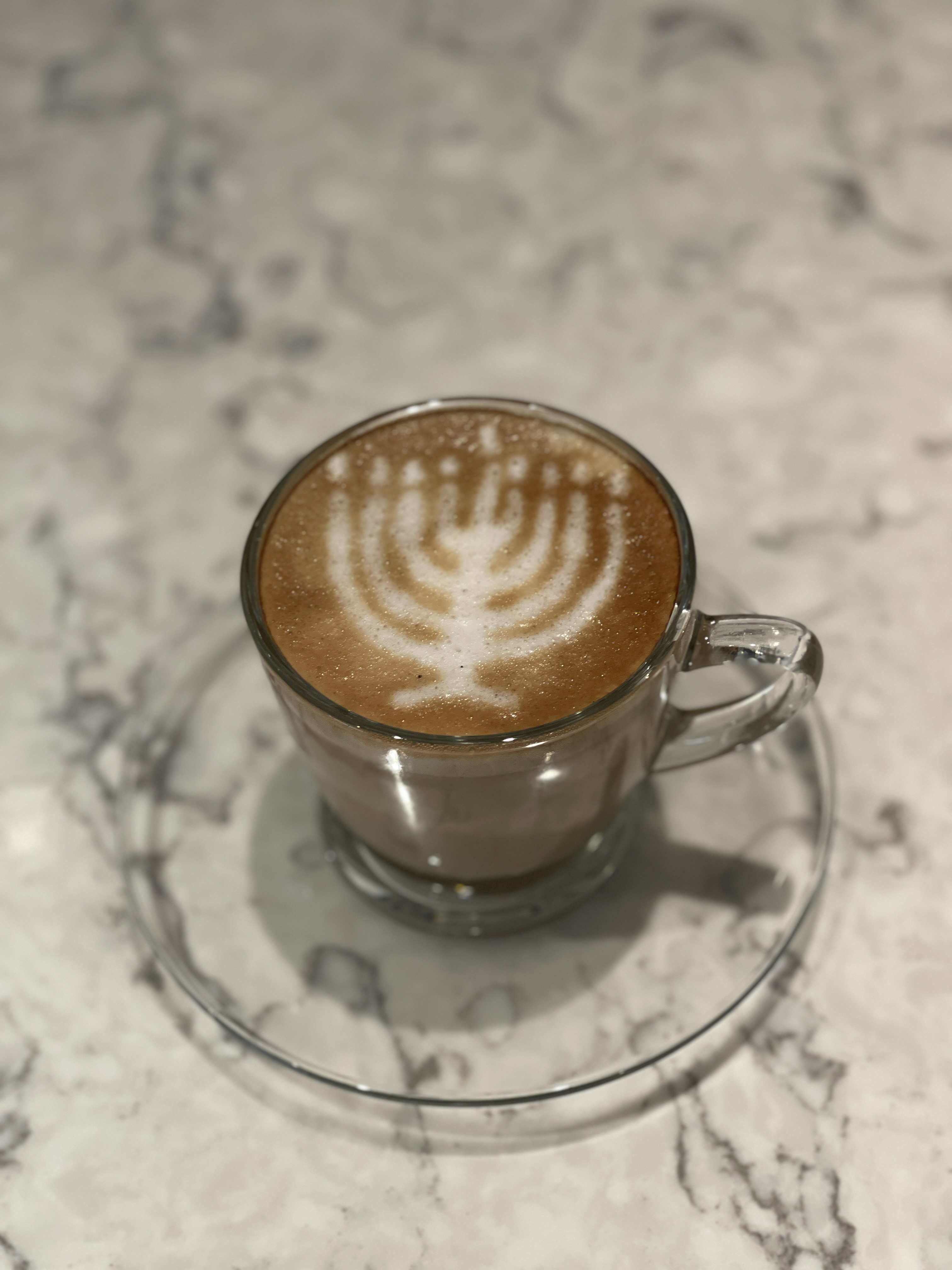 Cup of coffee with latte art shaped like a menorah on a clear glass saucer on a marble table.