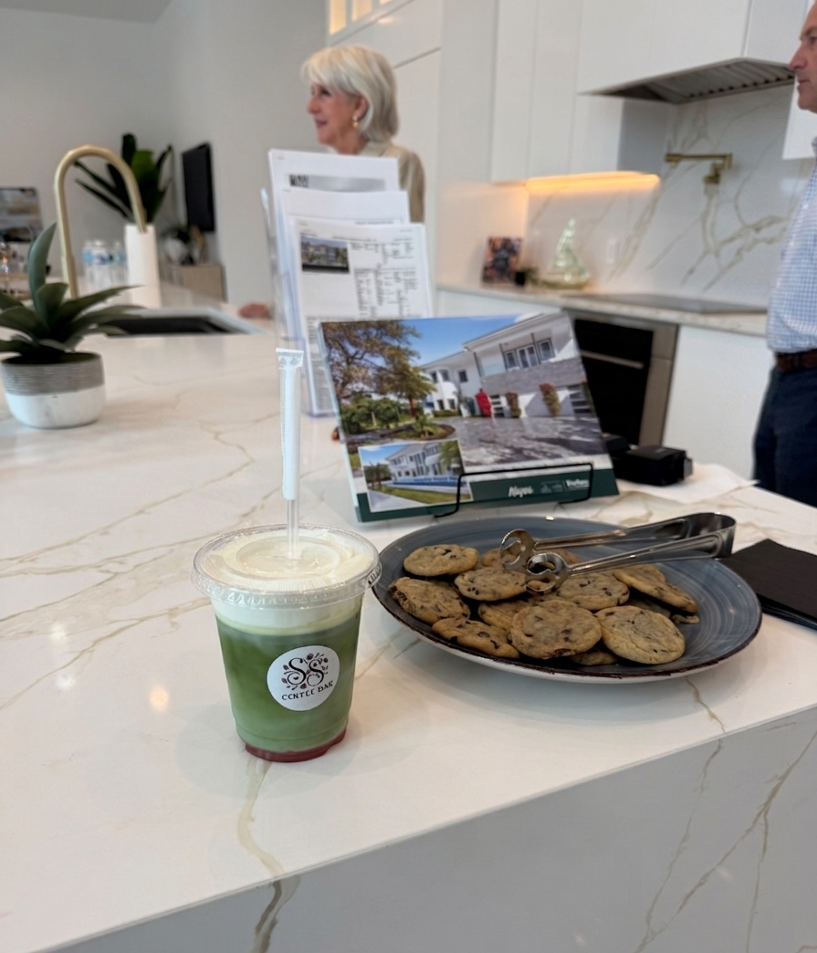 Plastic cup with layered green drink and lid next to a plate of chocolate chip cookies on a kitchen counter.