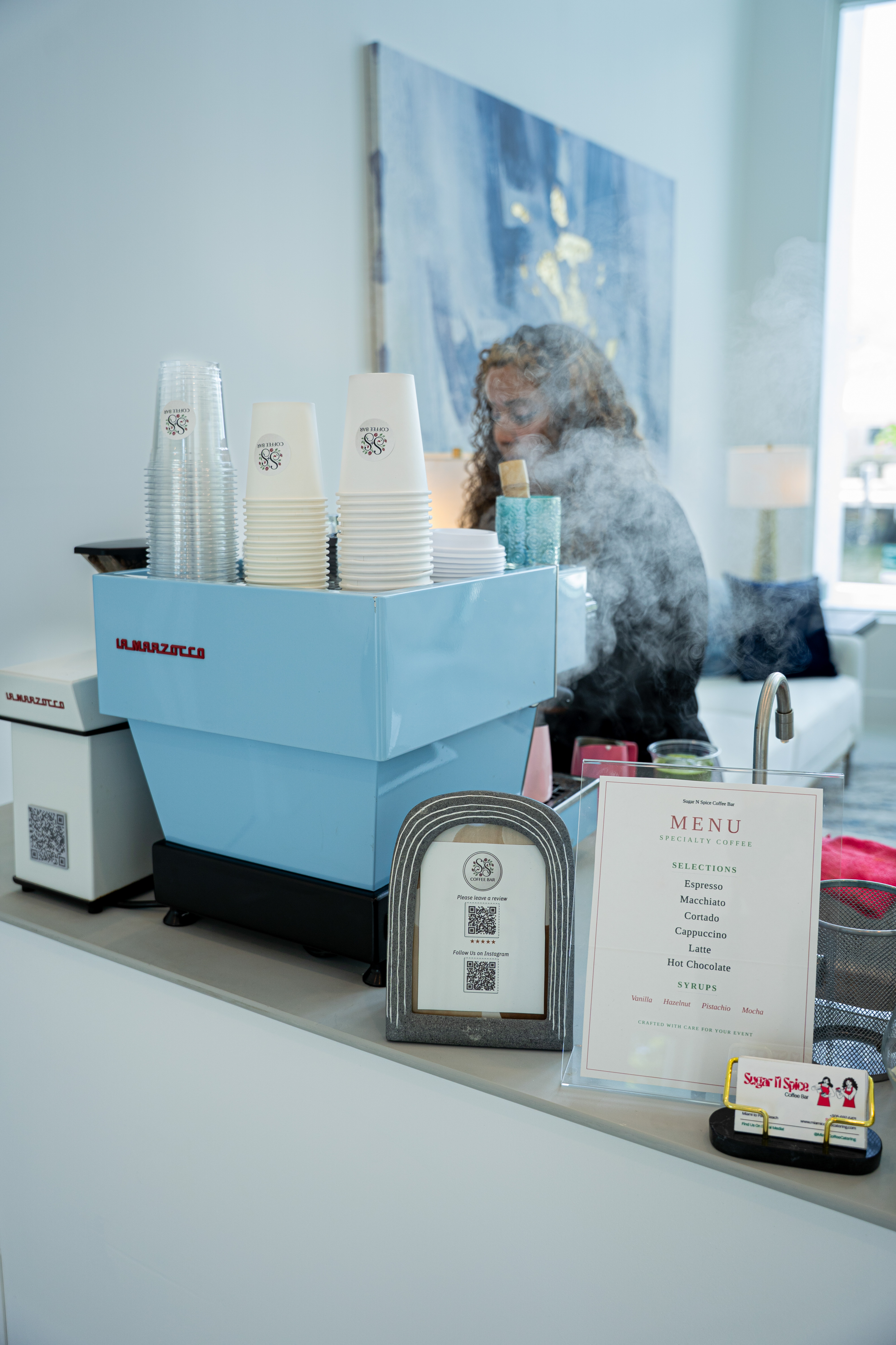 Blue espresso machine with stacked cups and a menu on a counter, barista working behind it.