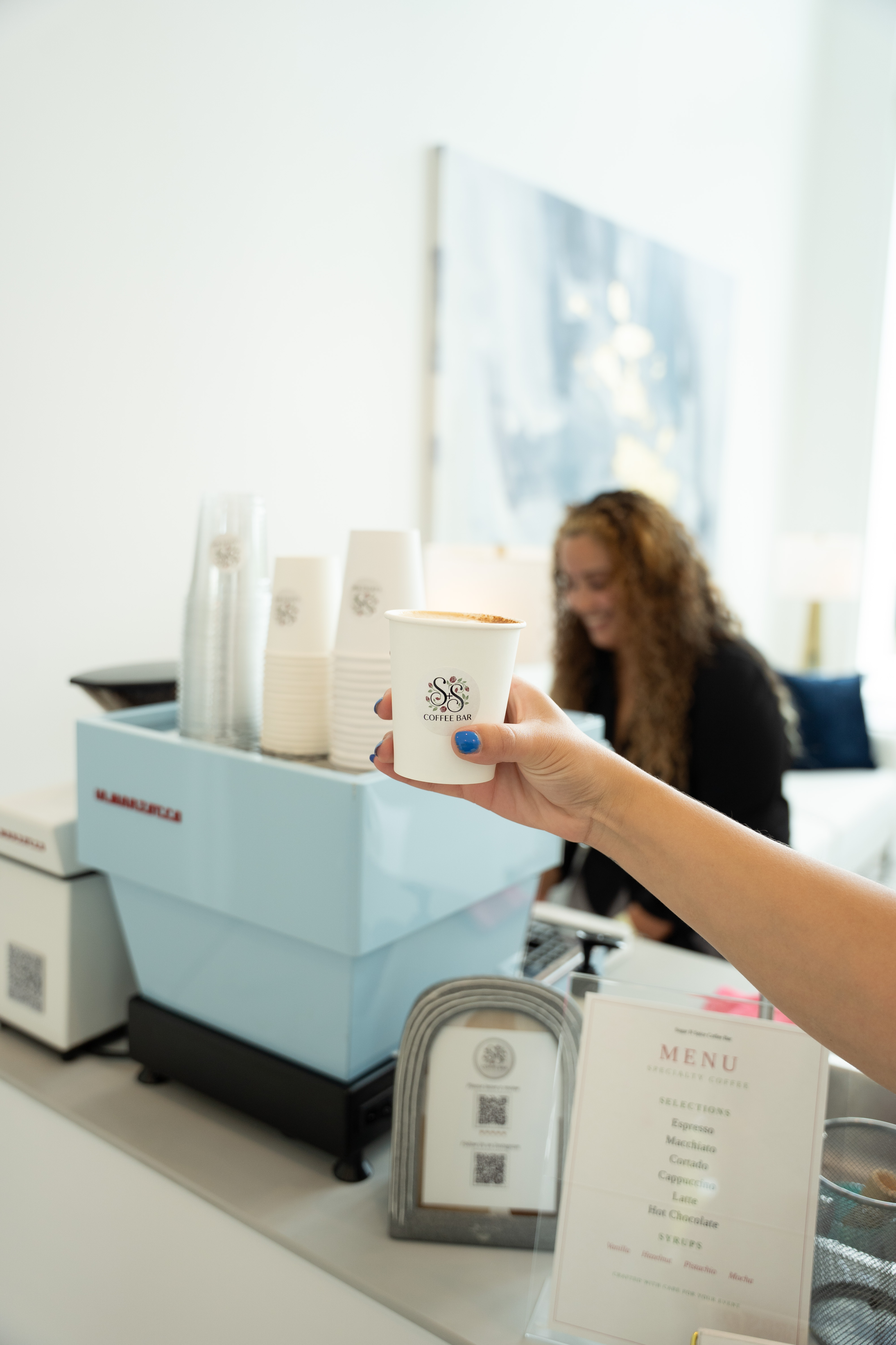 Hand with blue nail polish holding a coffee cup labeled SS Coffee Bar near a menu and a machine.