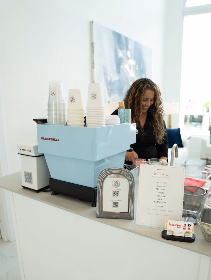 Coffee counter with a blue La Marzocco machine, stacked cups, menus, and a smiling barista in the background.
