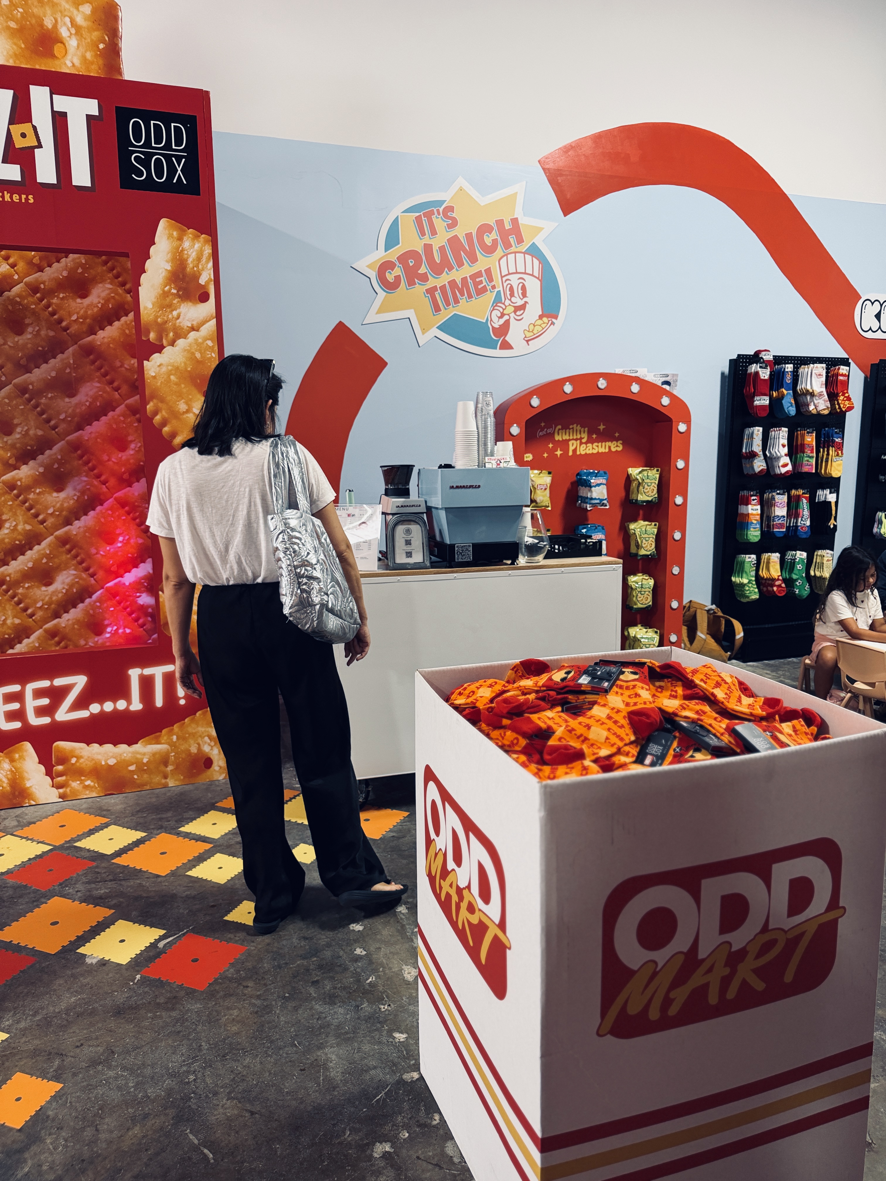 Woman standing inside a store near a large box of Odd Mart socks and a coffee counter.