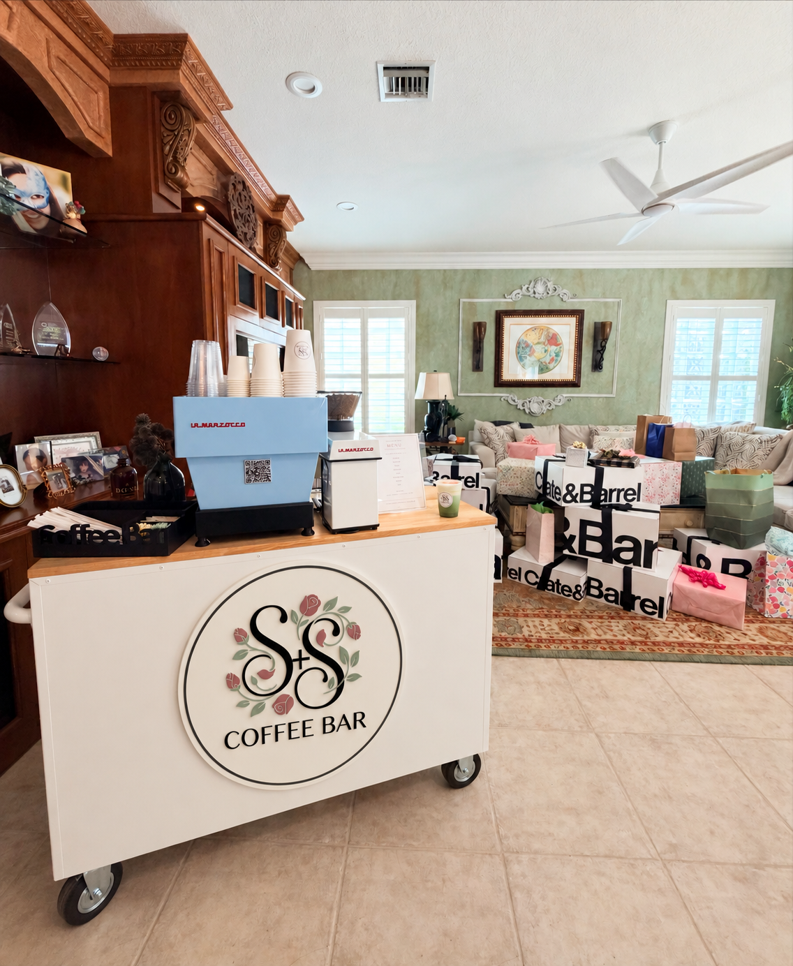 Indoor coffee bar cart with La Marzocco espresso machine, cups, and menu, set in a living room with a wooden cabinet and numerous Crate & Barrel gift bags on a couch and carpet.