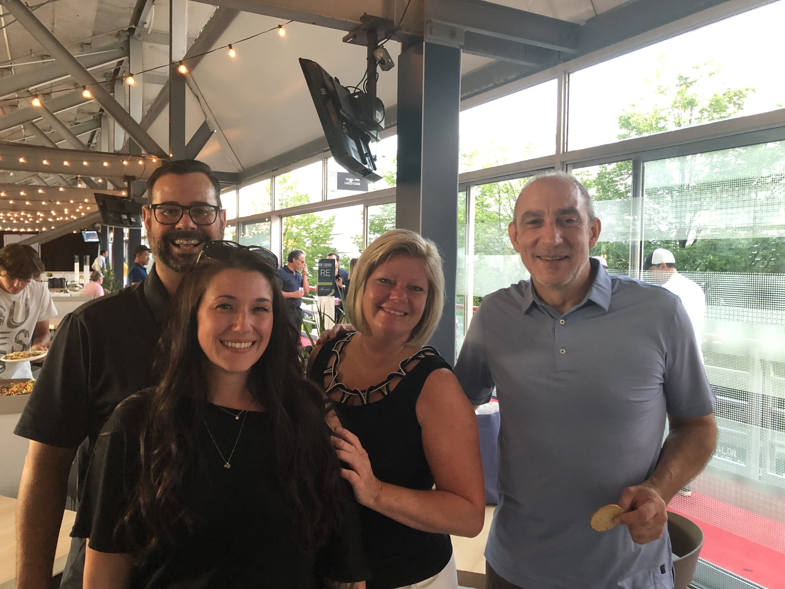 Four people smiling together at a bright, string-lit event space