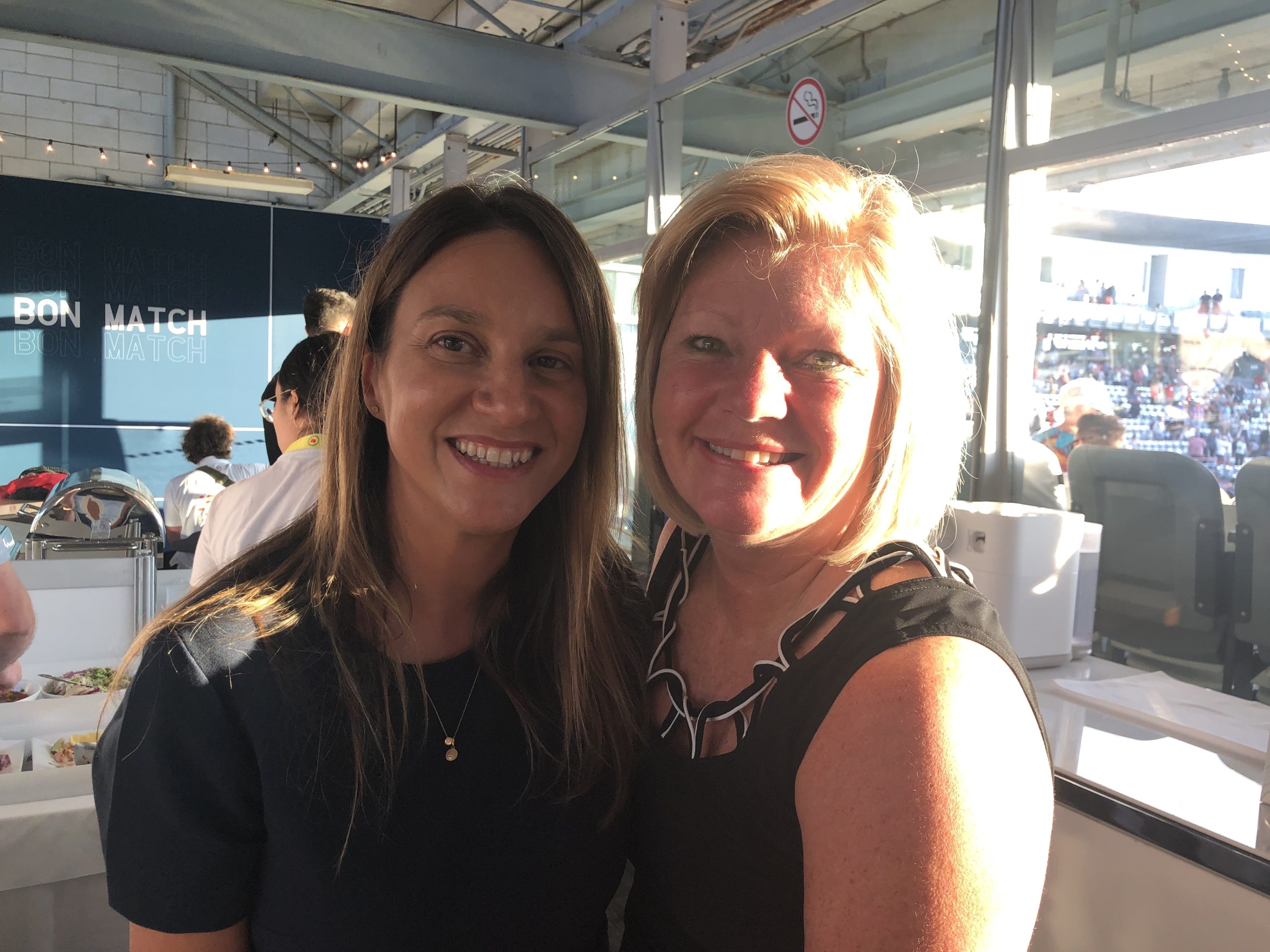 Two smiling women at a sports venue with 'Bon Match' sign
