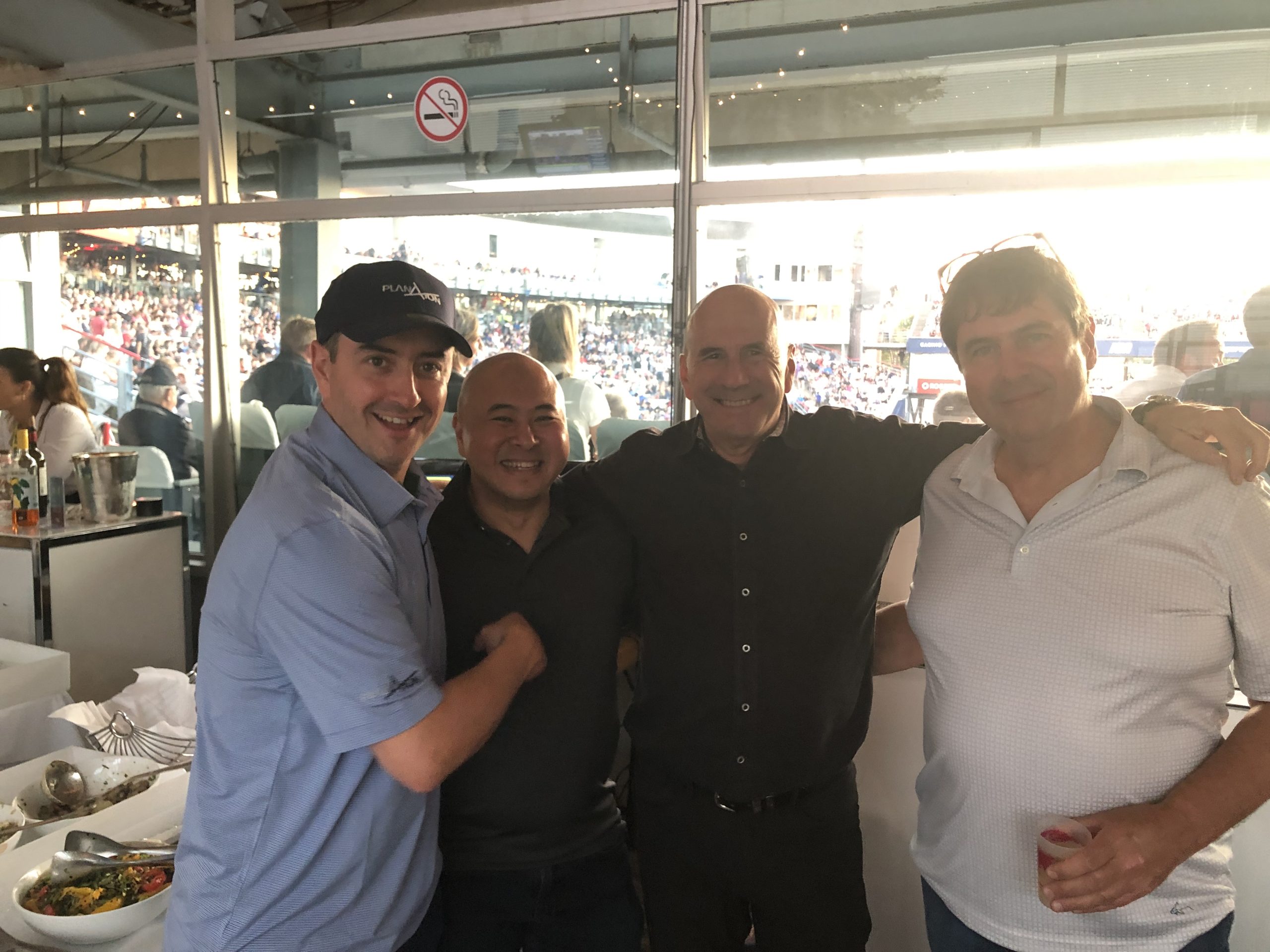 Four men smiling together at a sporting event with crowded stadium background
