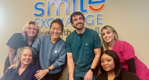 Group of six smiling dental professionals posing in front of a Smile Exchange sign.