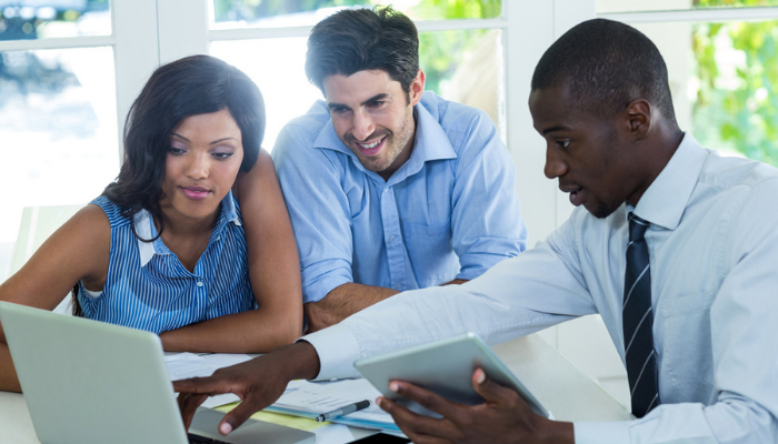 Three diverse professionals collaborating at a table with a laptop and tablet in a bright office.