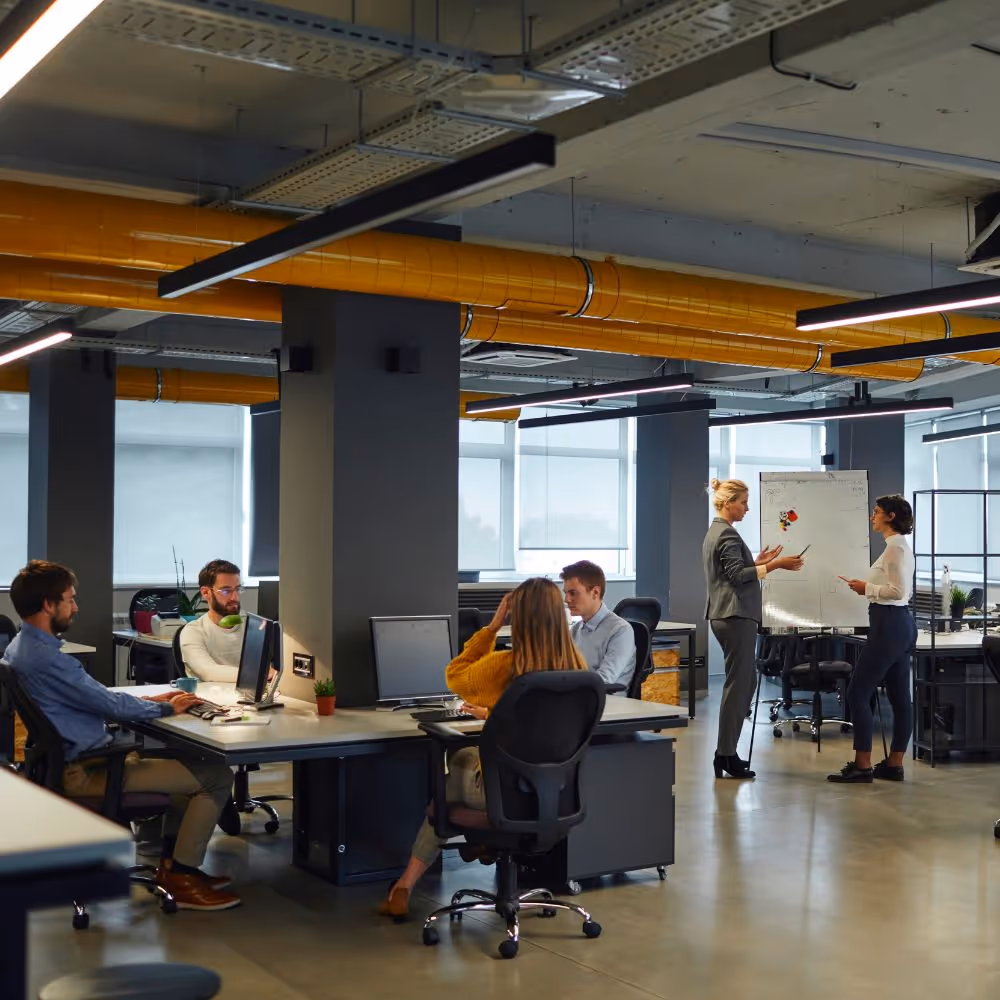 Modern office space with five people working; two women discussing at a whiteboard and three colleagues seated at desks with computers.