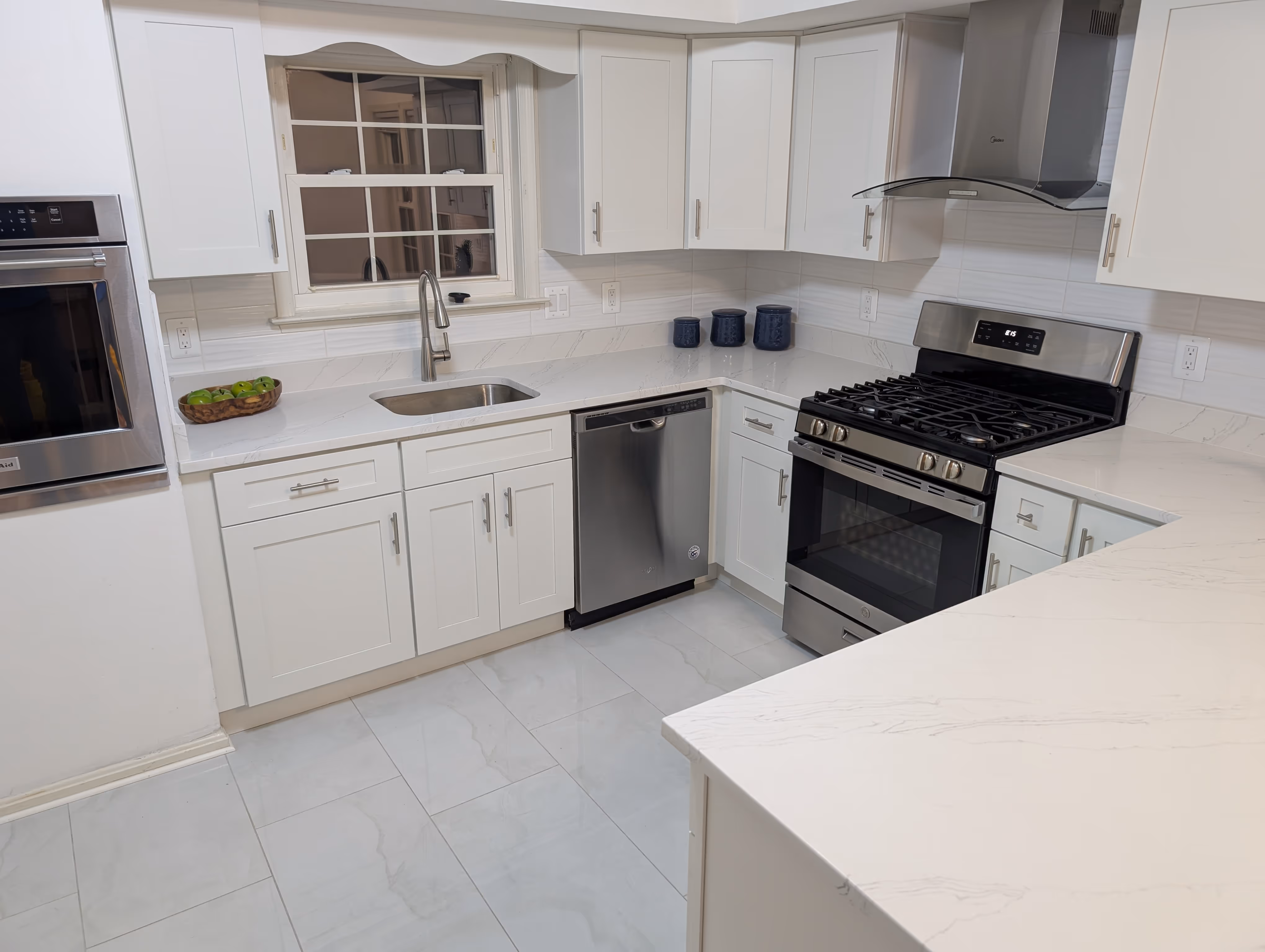 Modern kitchen with white cabinets, stainless steel appliances, a sink under a window, and a wooden bowl filled with green apples on the counter.