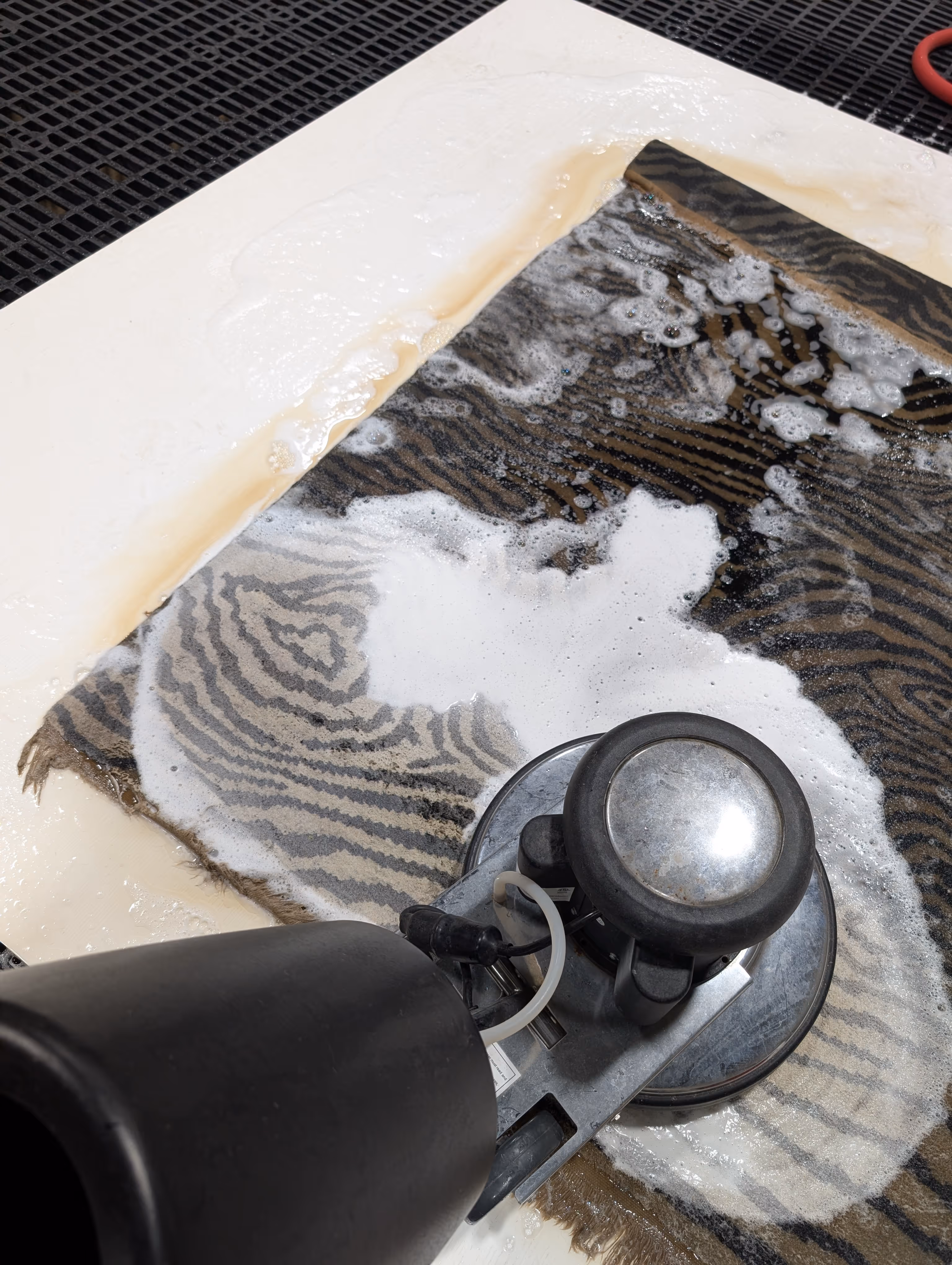 Rotary floor cleaning machine scrubbing a patterned rug with soap suds on a white floor near a metal grate.