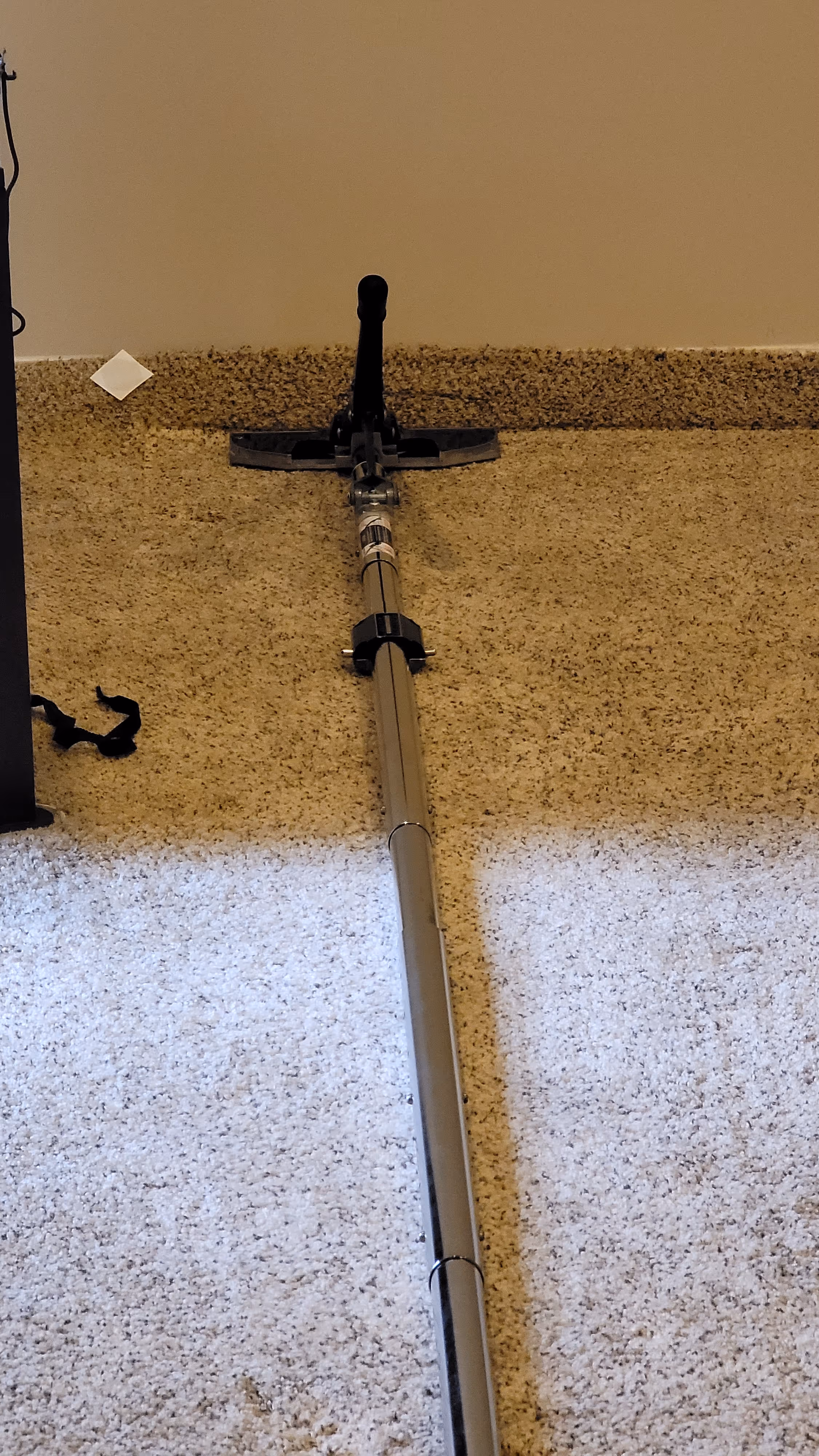 Long metal tool with a black handle and flat head resting on beige carpet against a tan wall.