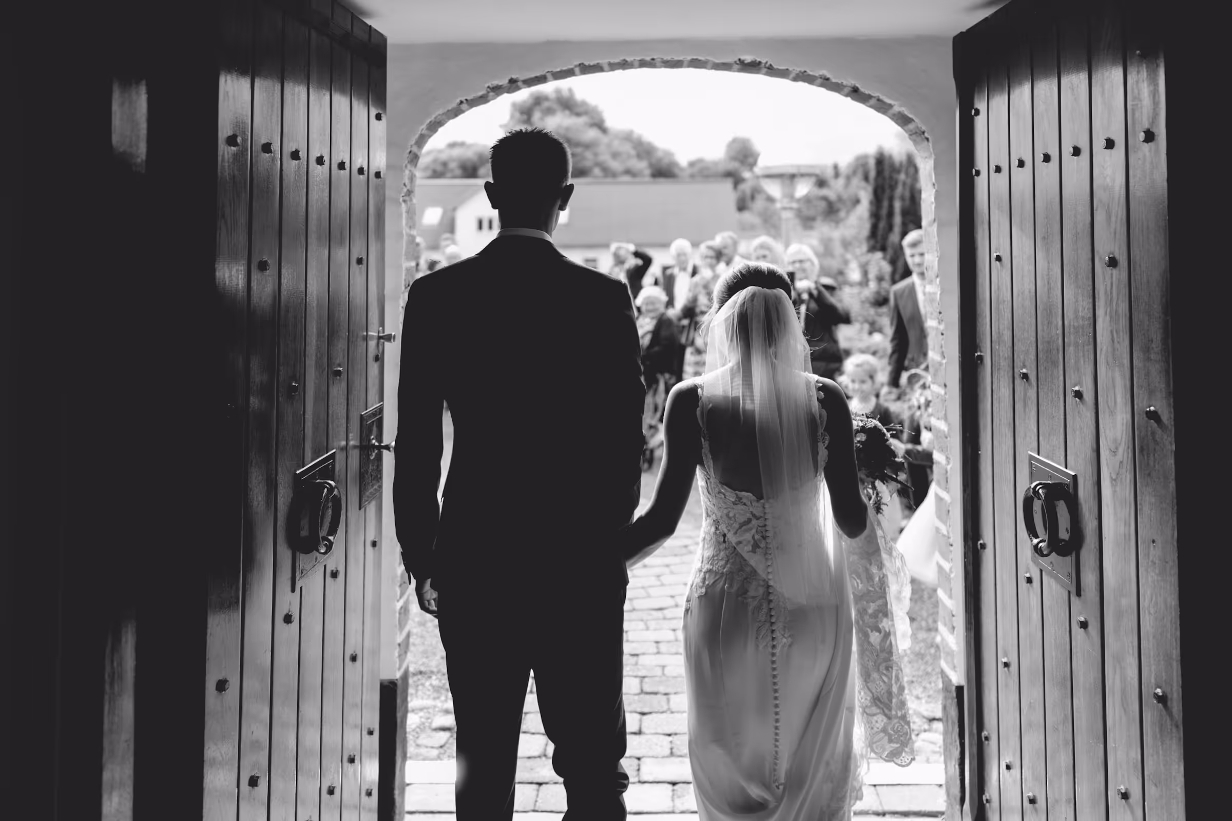 Back view of a bride and groom walking through open wooden doors toward their wedding guests outside.