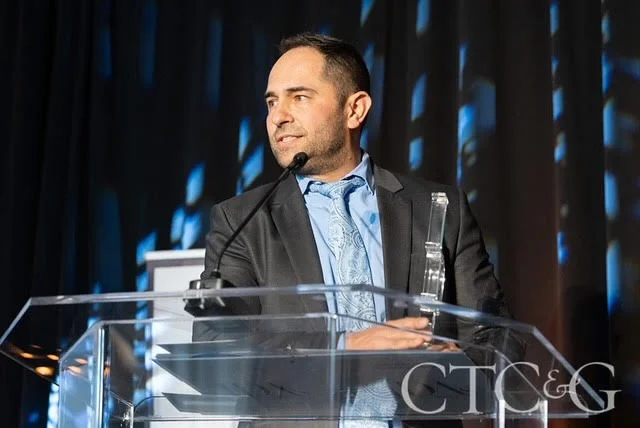 A man in a suit speaking at a podium while holding an award trophy.
