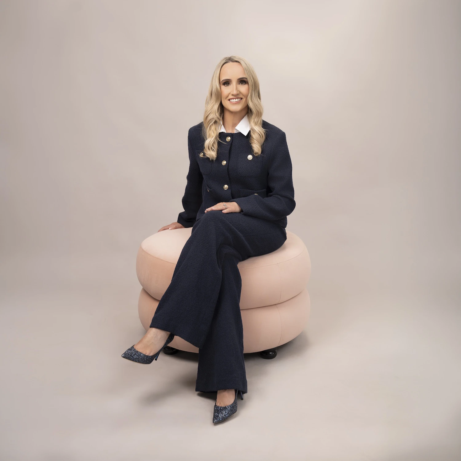 Blonde woman in navy suit with gold buttons sitting on a peach ottoman, smiling at camera.