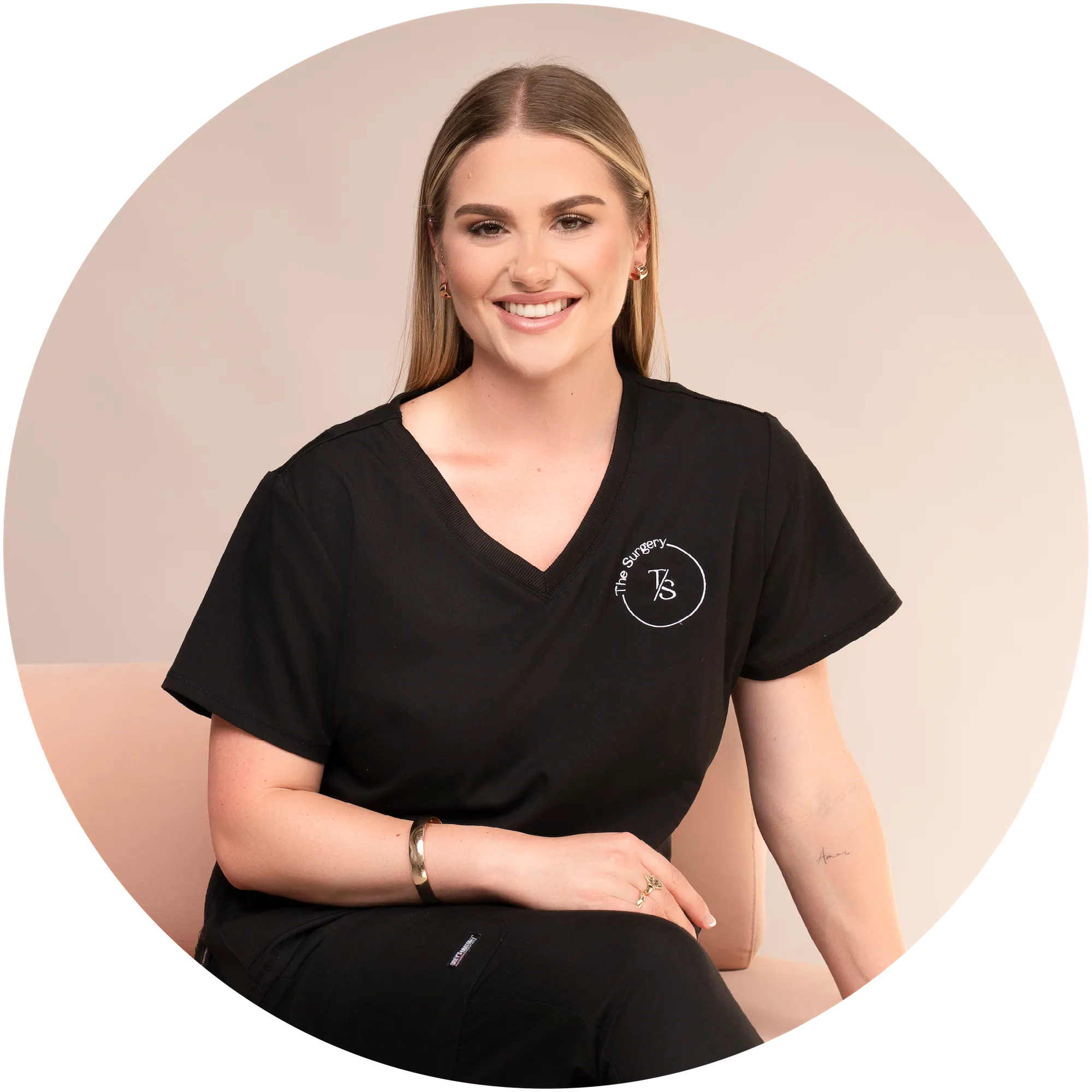 Smiling woman with long blonde hair in black scrubs sitting on a beige chair.