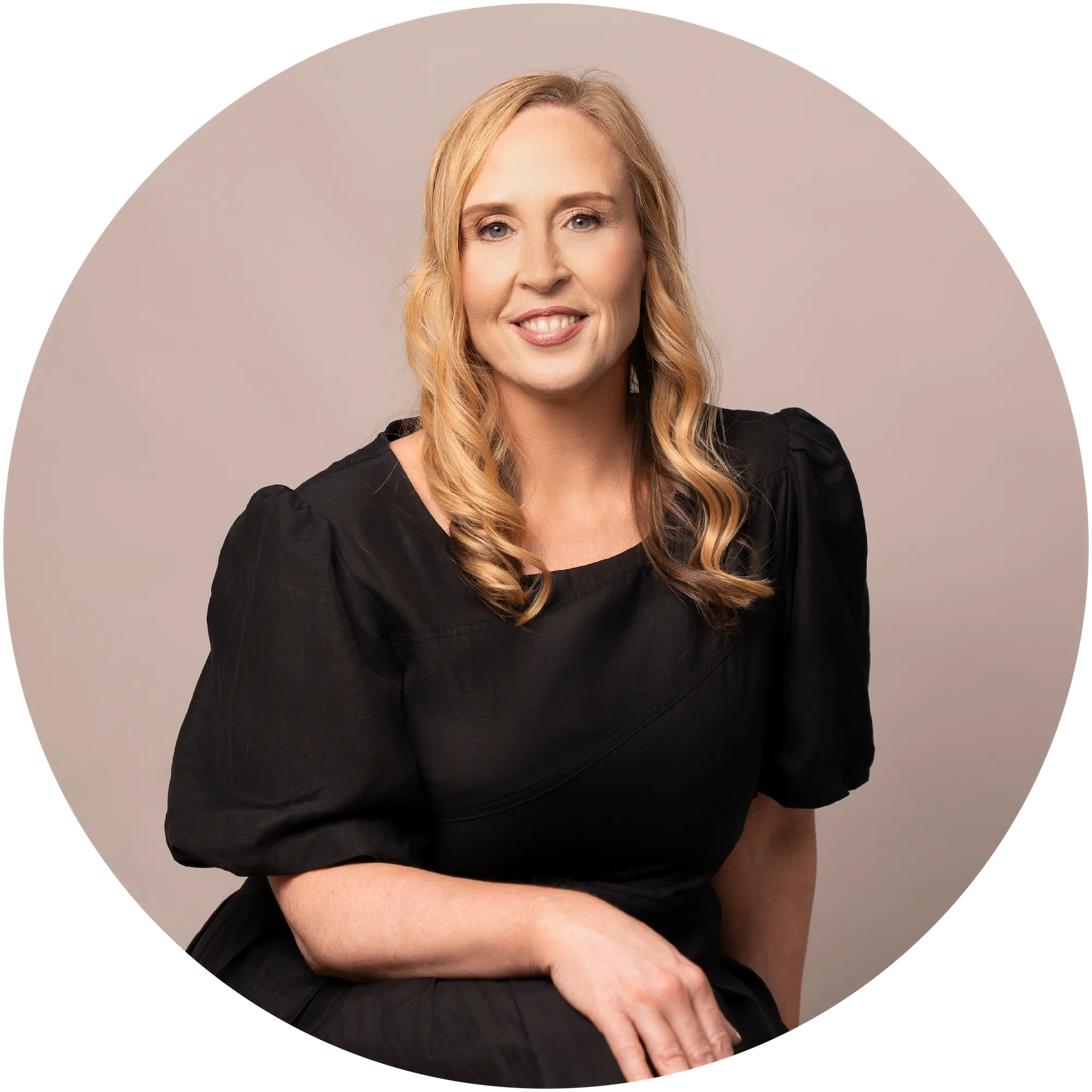 Smiling woman with curled blonde hair wearing a black dress, seated against a beige background.