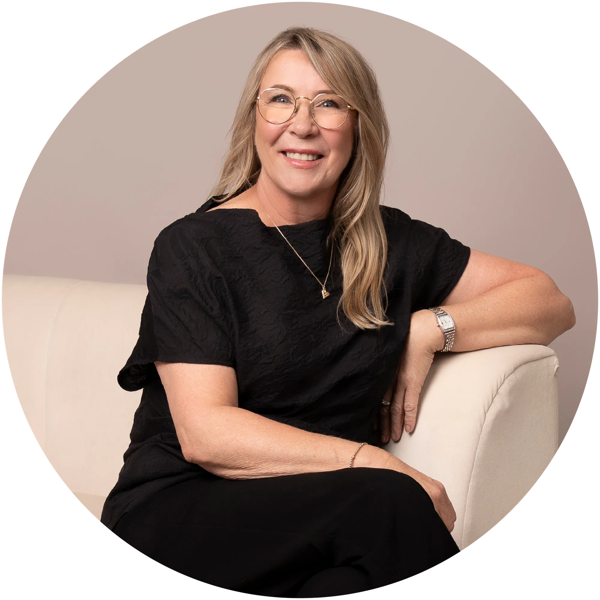 Smiling middle-aged woman in glasses and black shirt sitting on a cream sofa against a beige background.