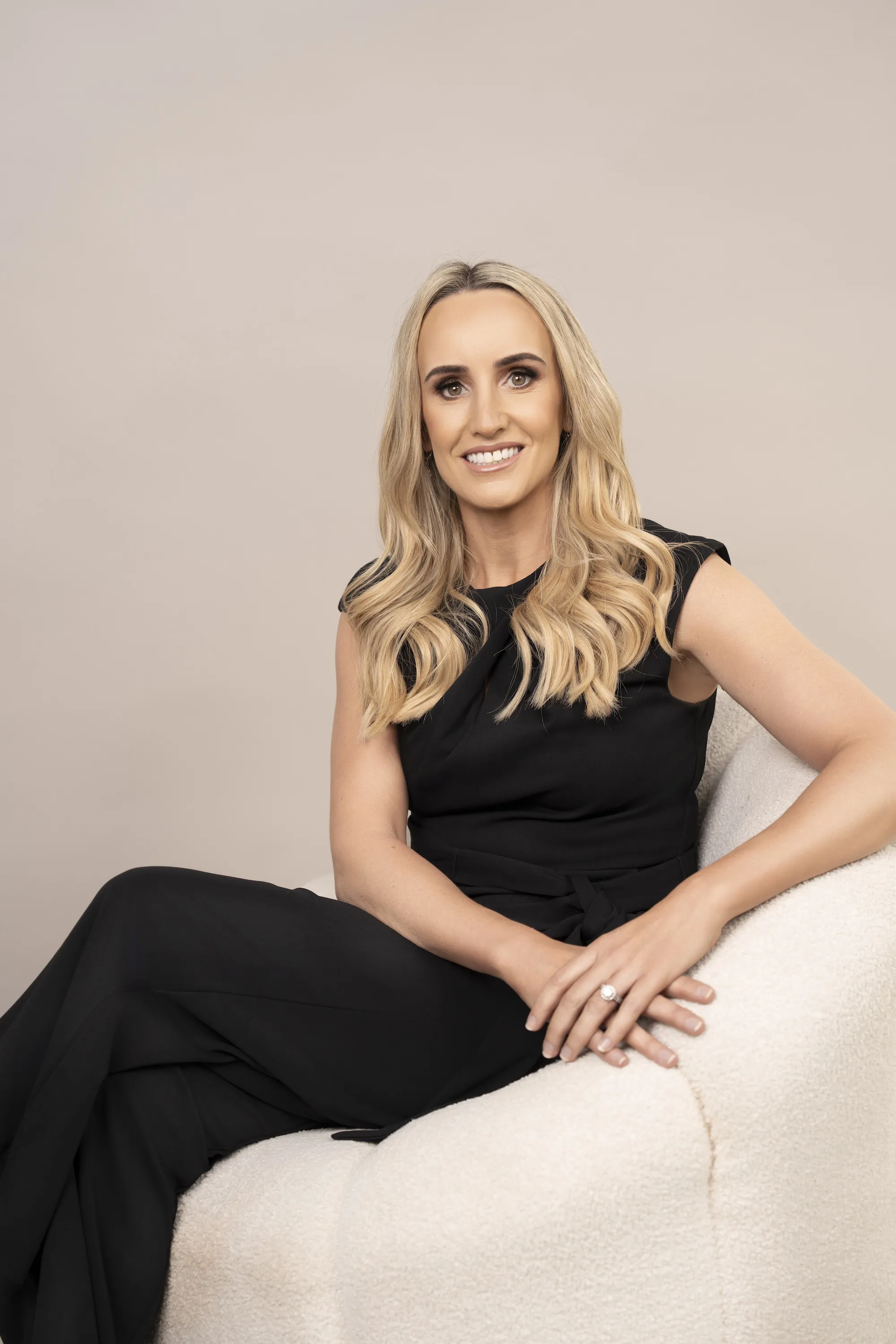 Smiling woman with long blonde hair wearing a black dress sitting on a white chair.