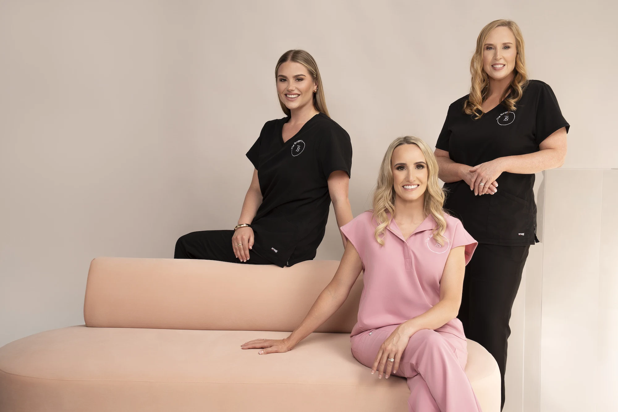 Three women smiling, two in black uniforms and one in pink, posing on and beside a peach couch.