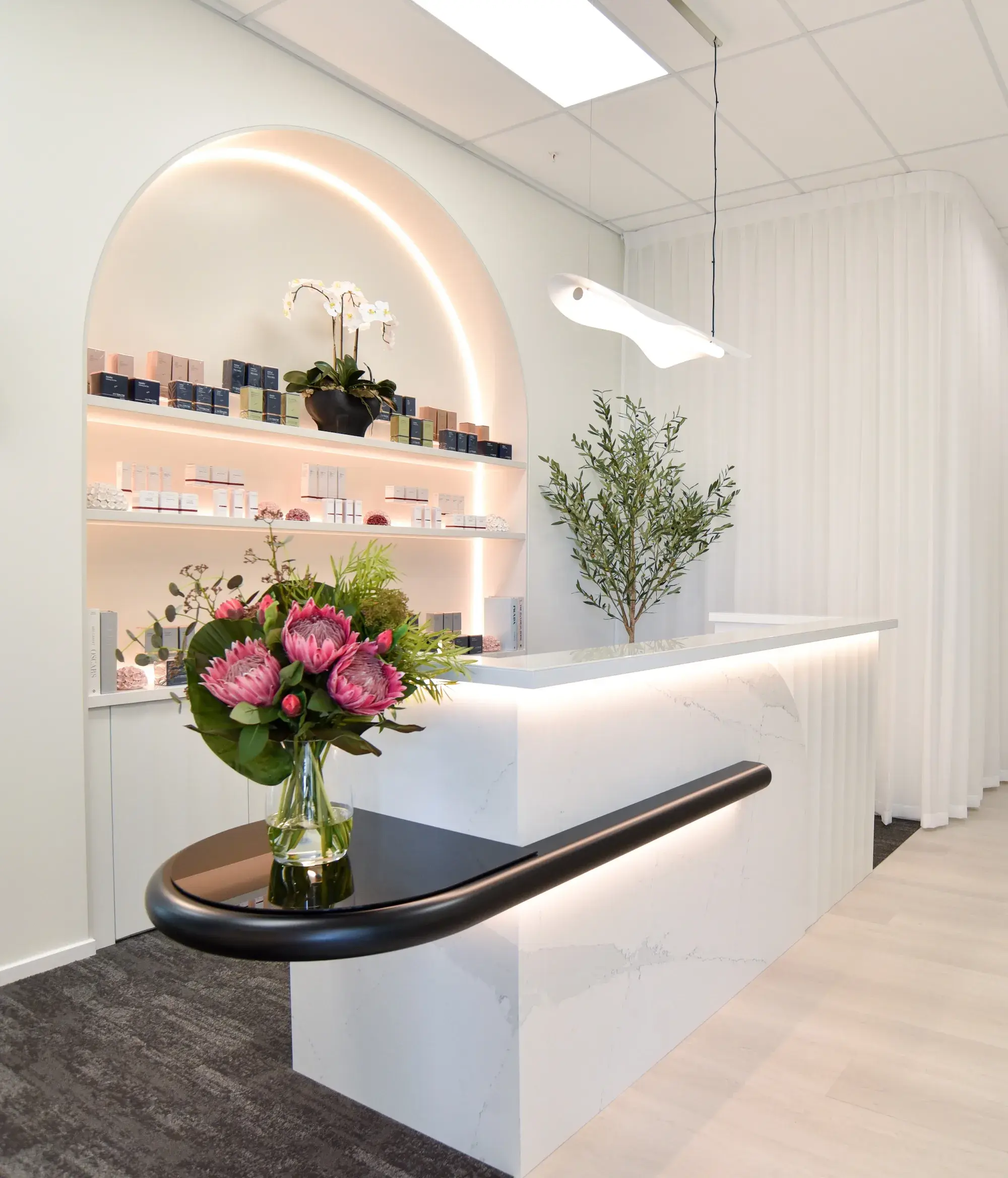 Modern white reception desk with black ledge, floral bouquet, backlit shelves, and hanging light.