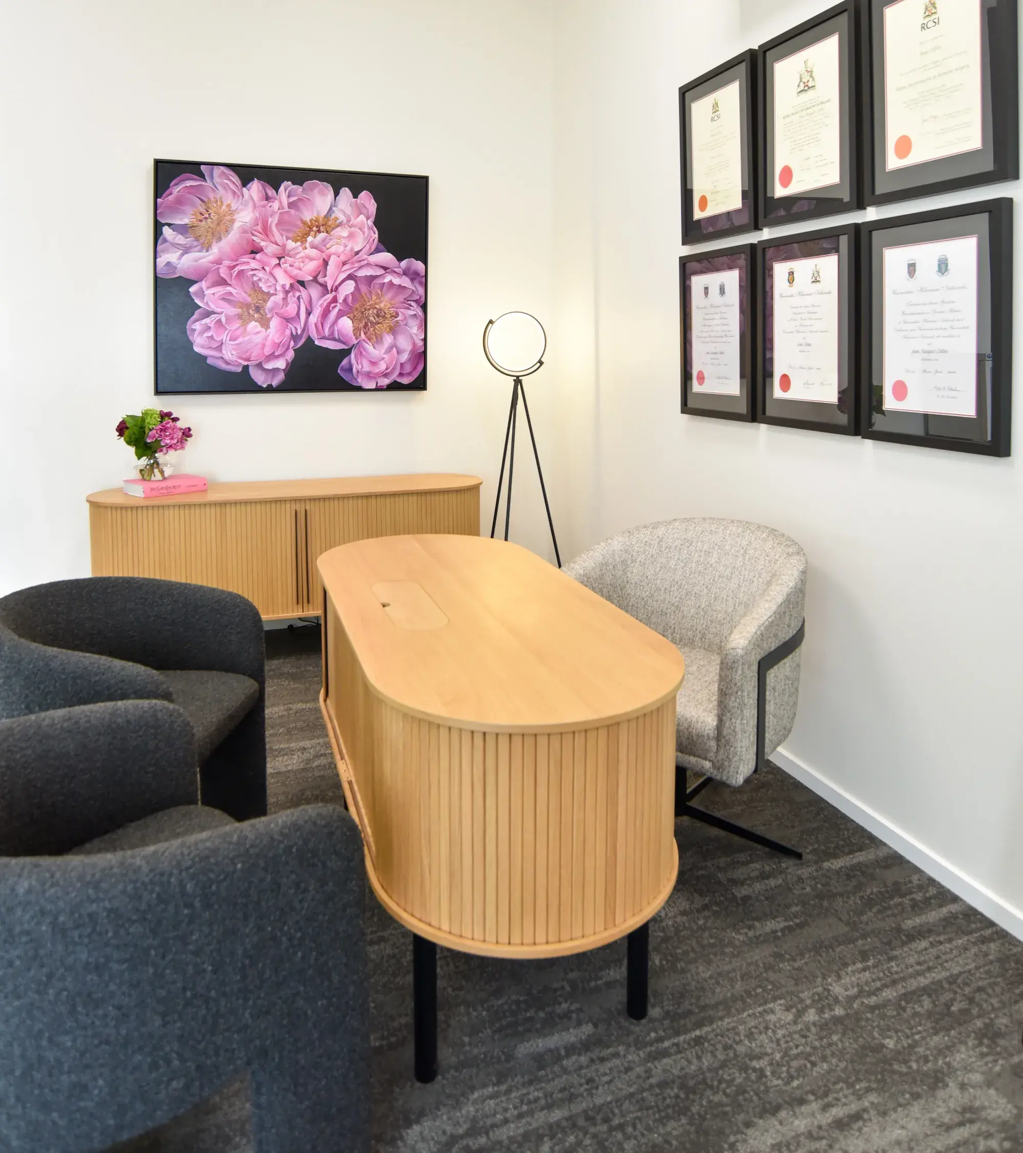 Modern office with wooden desk, gray and beige chairs, floral painting, certificates on wall, and floor lamp.