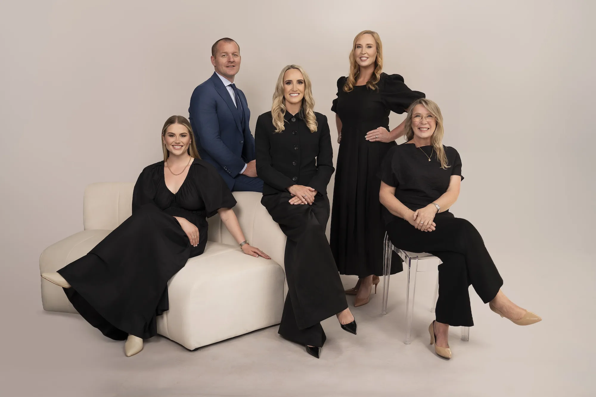 Five professionally dressed people posed in a studio with a neutral background and light seating.