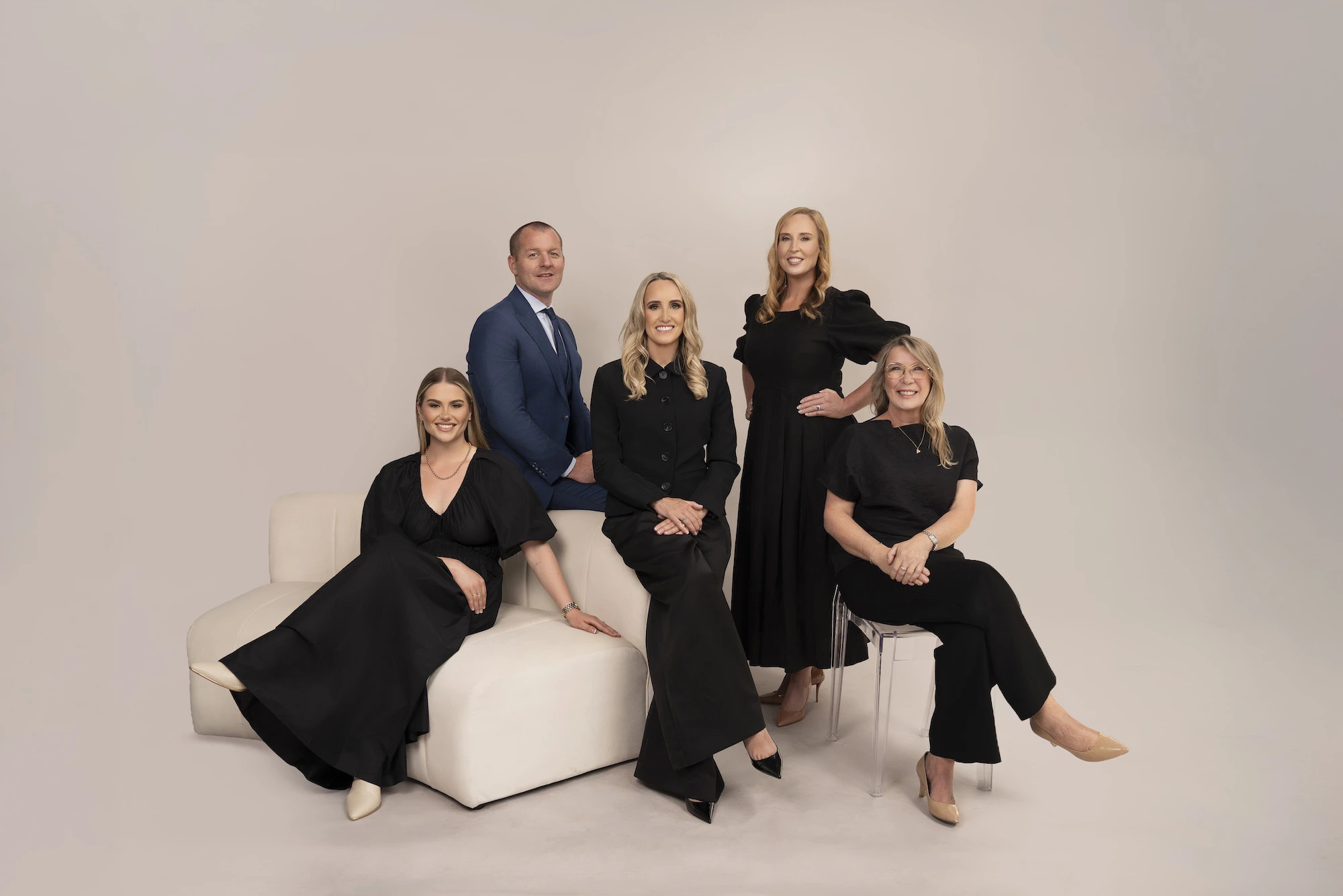 Group of four women in black outfits and one man in a blue suit posing in a studio with neutral backdrop.